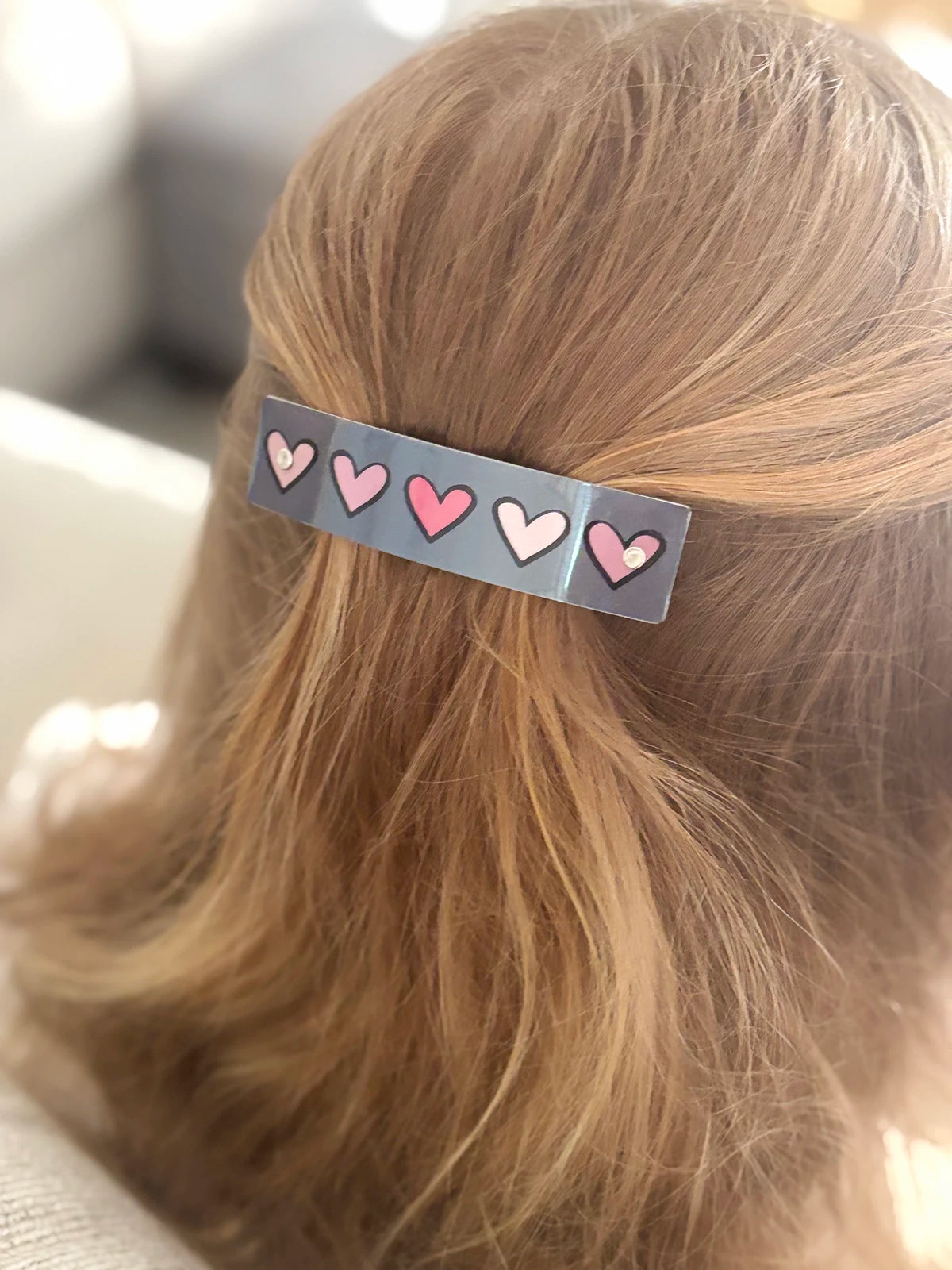 A close-up of a person's blonde hair secured with a gray hair clip decorated with pink hearts.