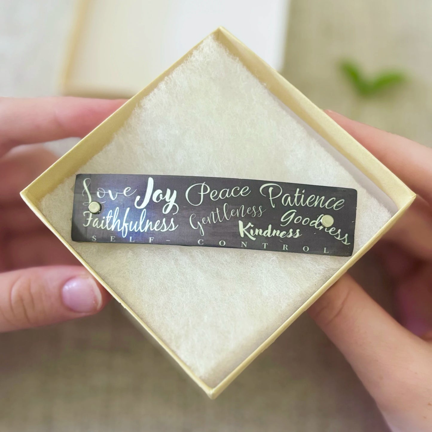 A rectangular gray hair barrette with silver script text reading 'Love Joy Peace Patience Faithfulness Gentleness Goodness Kindness SELF CONTROL' is held in a person's hands inside a square cream-colored gift box with a textured insert.