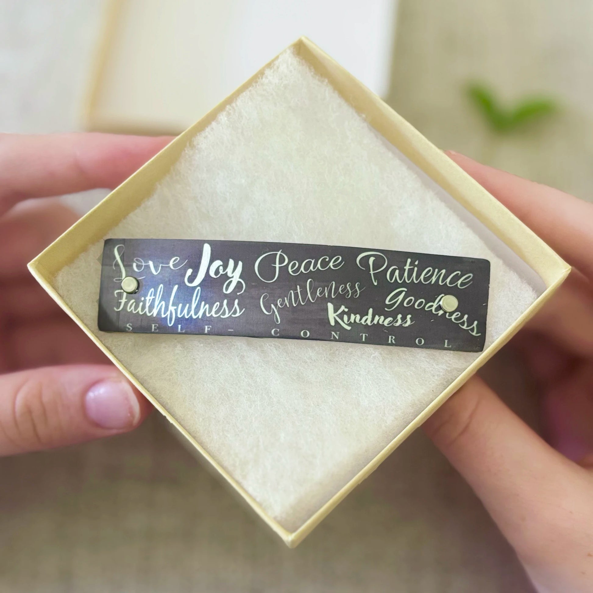 A rectangular gray hair barrette with silver script text reading 'Love Joy Peace Patience Faithfulness Gentleness Goodness Kindness SELF CONTROL' is held in a person's hands inside a square cream-colored gift box with a textured insert.