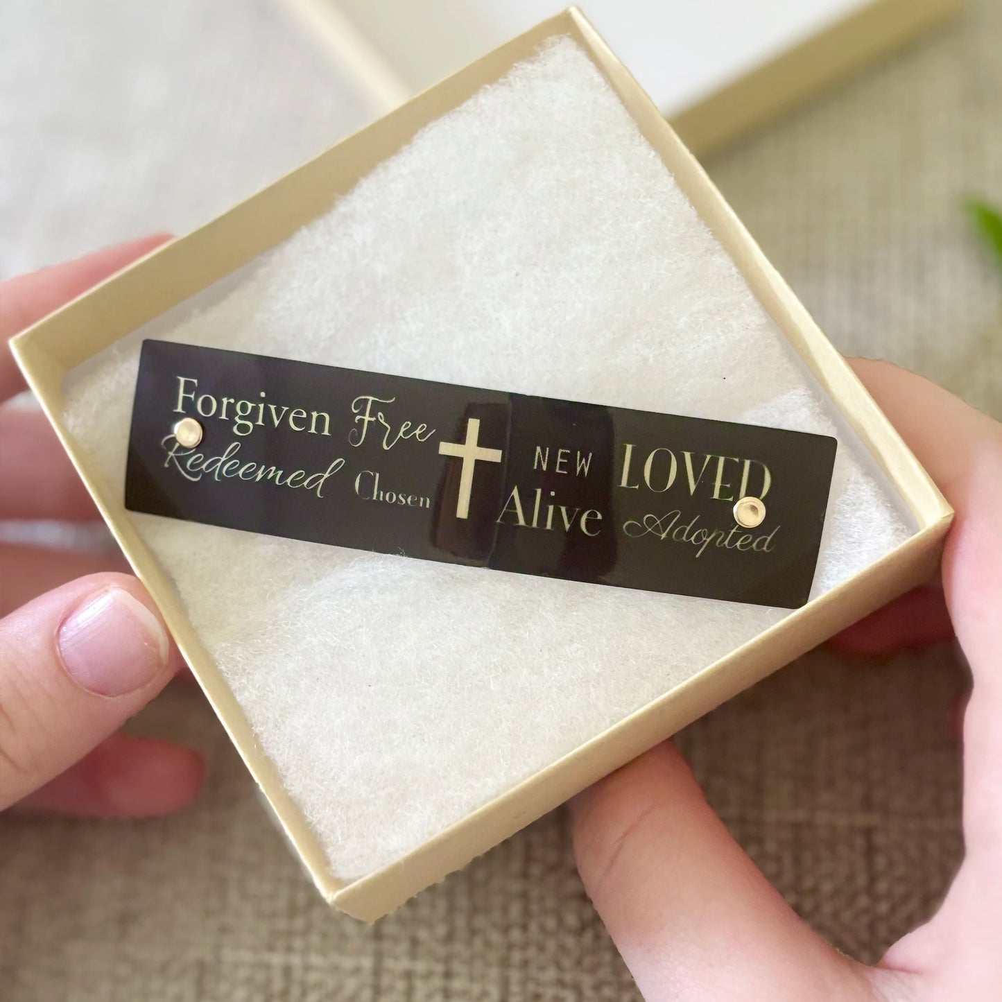 A close-up of a person holding a cream-colored square gift box with a black barrette that reads 'Forgiven Free Redeemed Chosen New Alive Loved Adopted' with a cross in the center. The person's hands are visible holding the box against a neutral textured background.