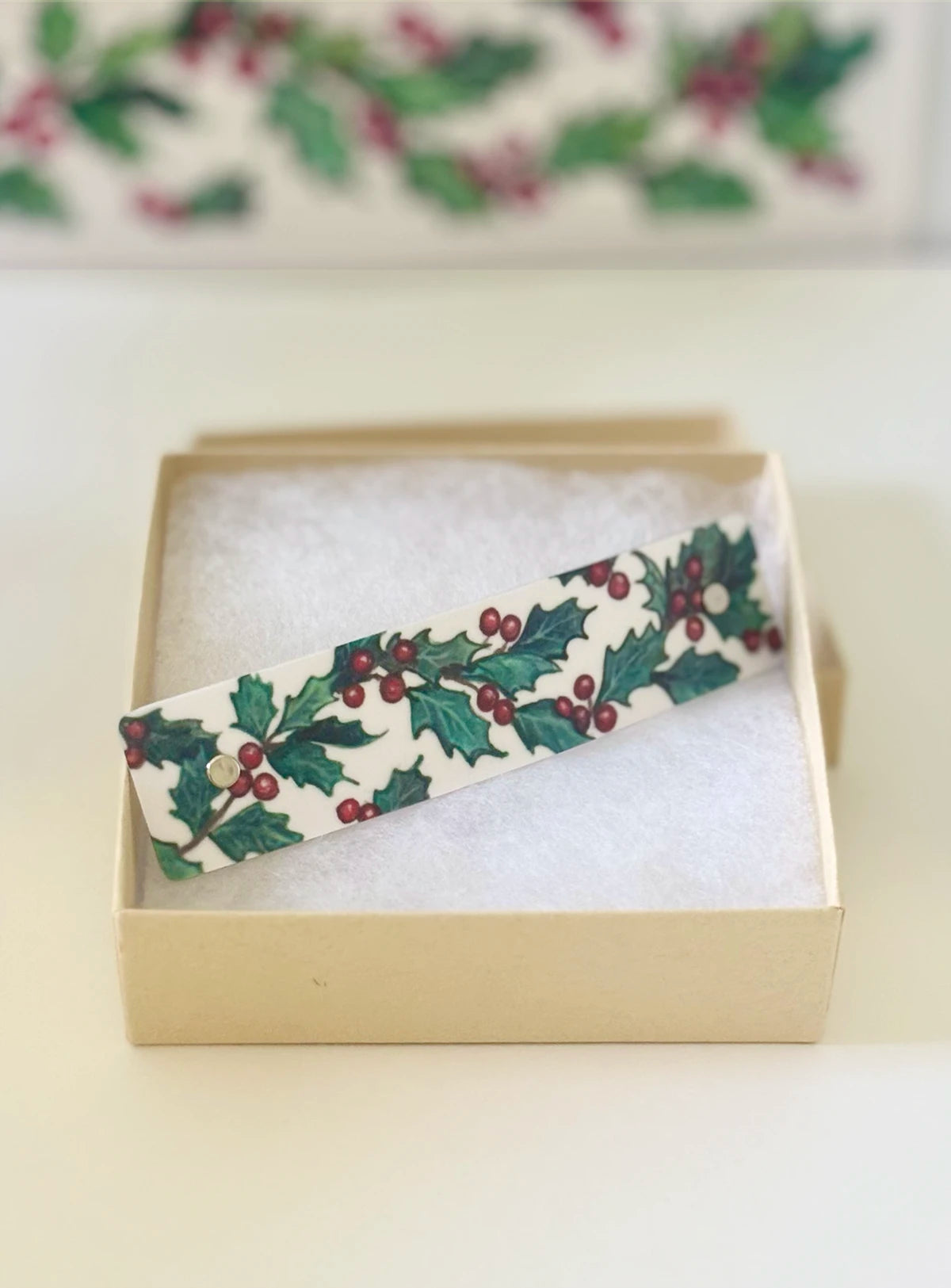 A rectangular white hair clip with a green holly and red berry pattern, resting inside an open beige gift box.