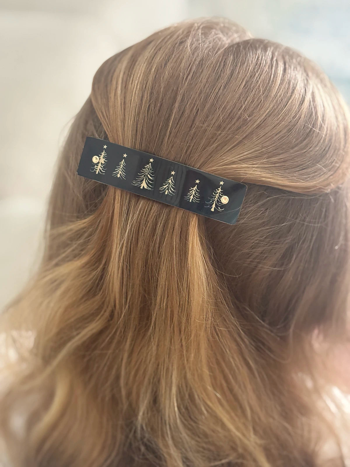 A close-up of a person's light brown hair secured with a black metal hair clip decorated with small gold Christmas tree and star motifs.