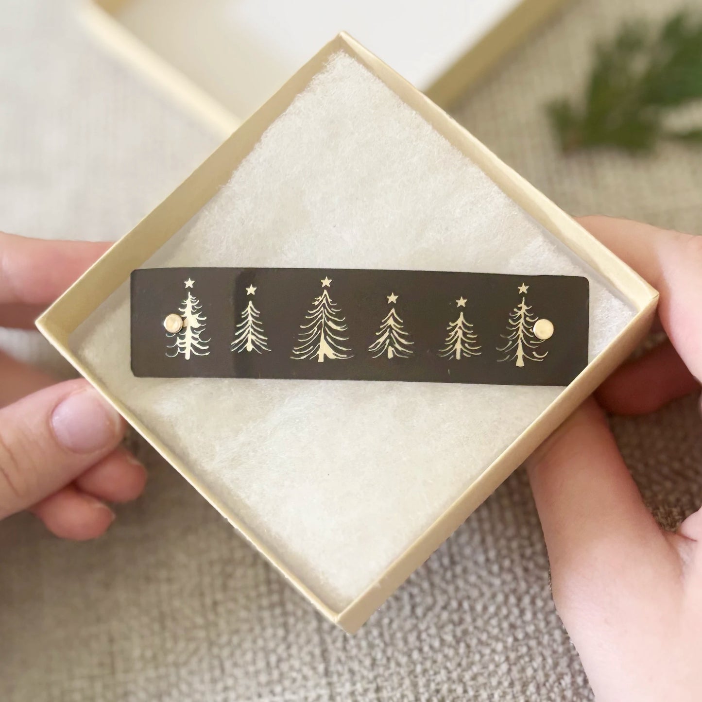 A close-up of hands holding an open beige gift box containing a black hair clip with gold Christmas tree and star designs, set against a soft neutral background.