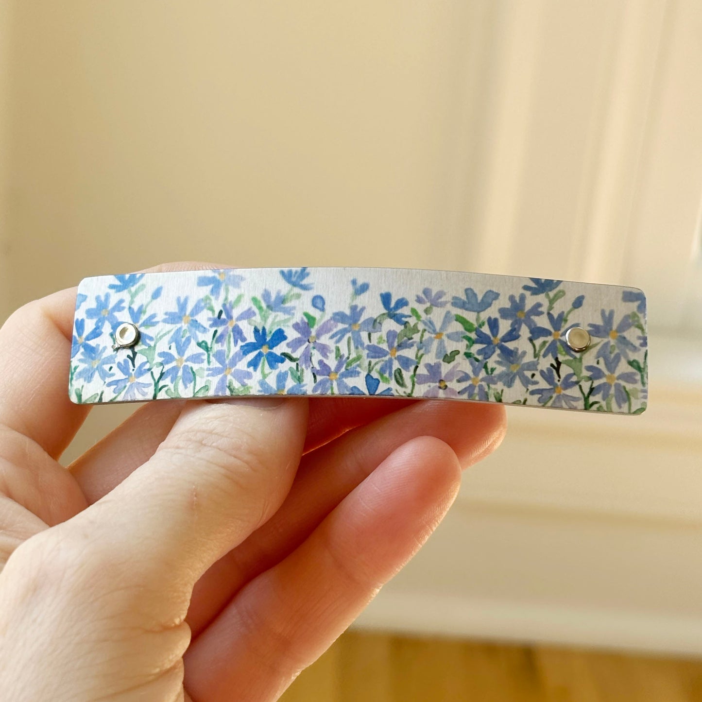 A close-up of a hand holding a slim rectangular hair clip decorated with a blue and lavender floral pattern on a silver metal background.