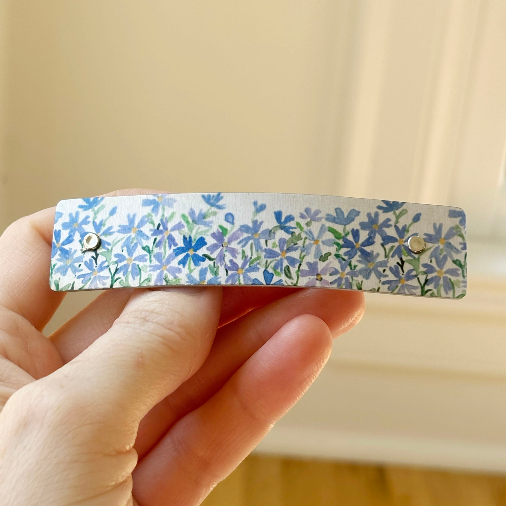 A close-up of a hand holding a slim rectangular hair clip decorated with a blue and lavender floral pattern on a silver metal background.