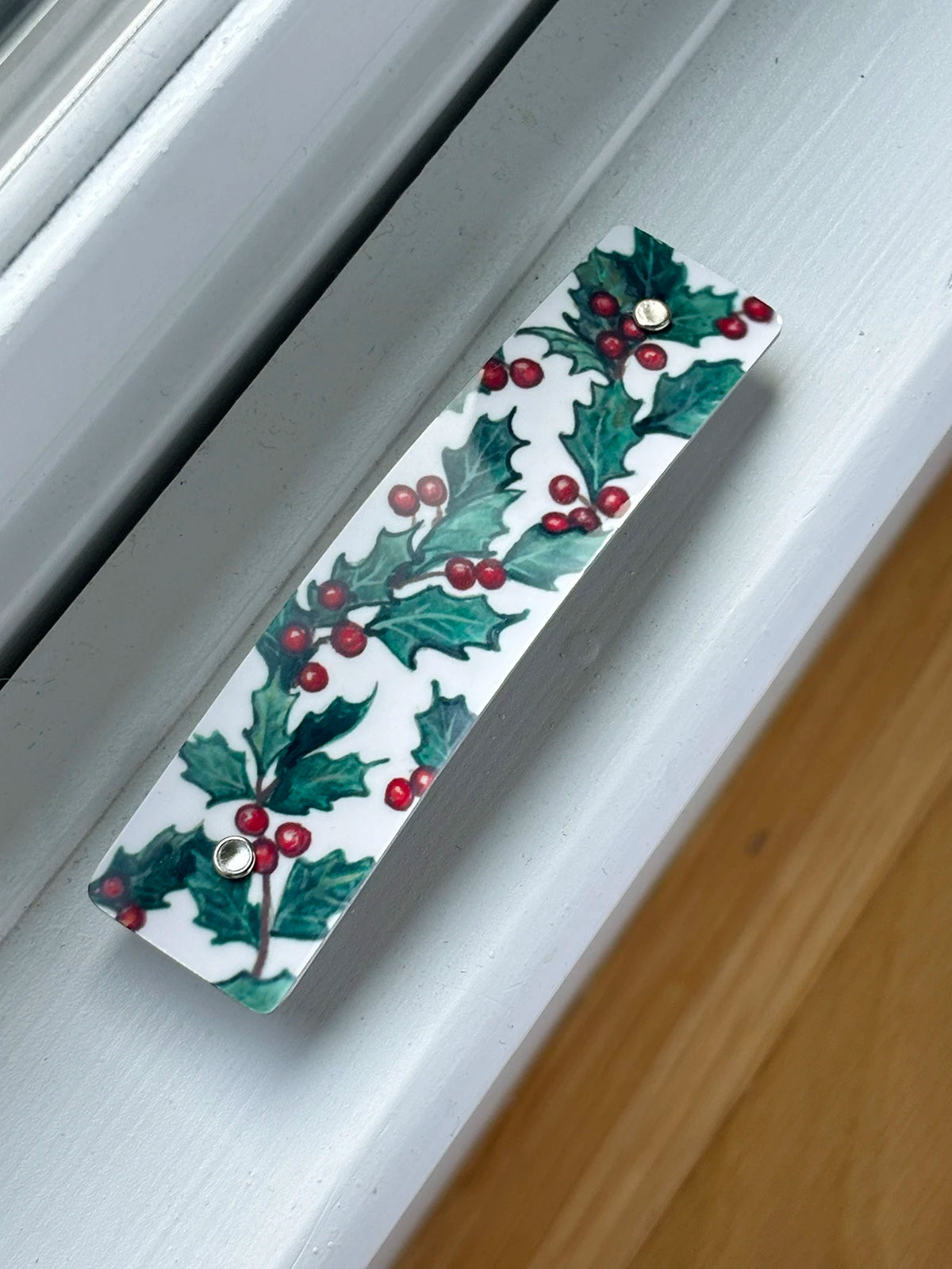 A rectangular white hair French clip decorated with a green holly and red berry pattern, shown resting on a white window sill with a wooden floor visible in the corner.