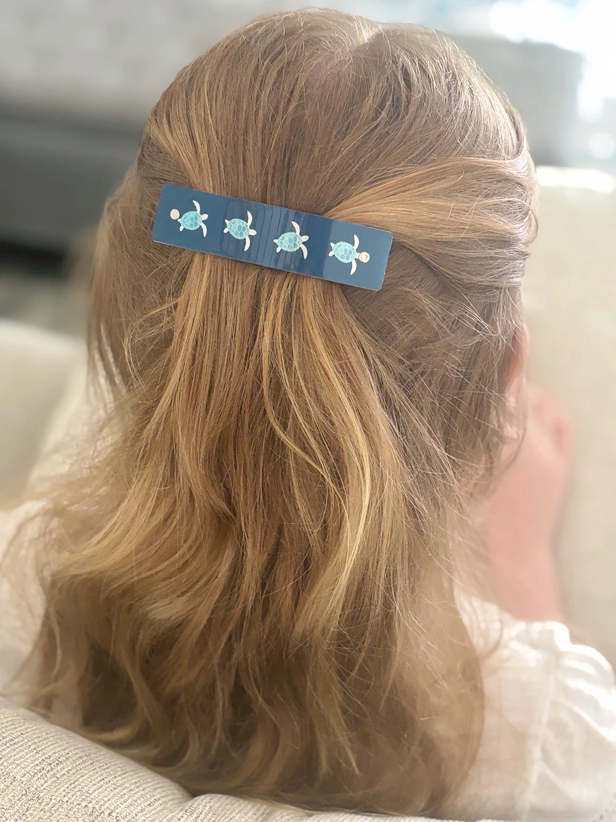 A close-up back view of a person with blond hair wearing a blue hair clip decorated with four small sea turtle illustrations.