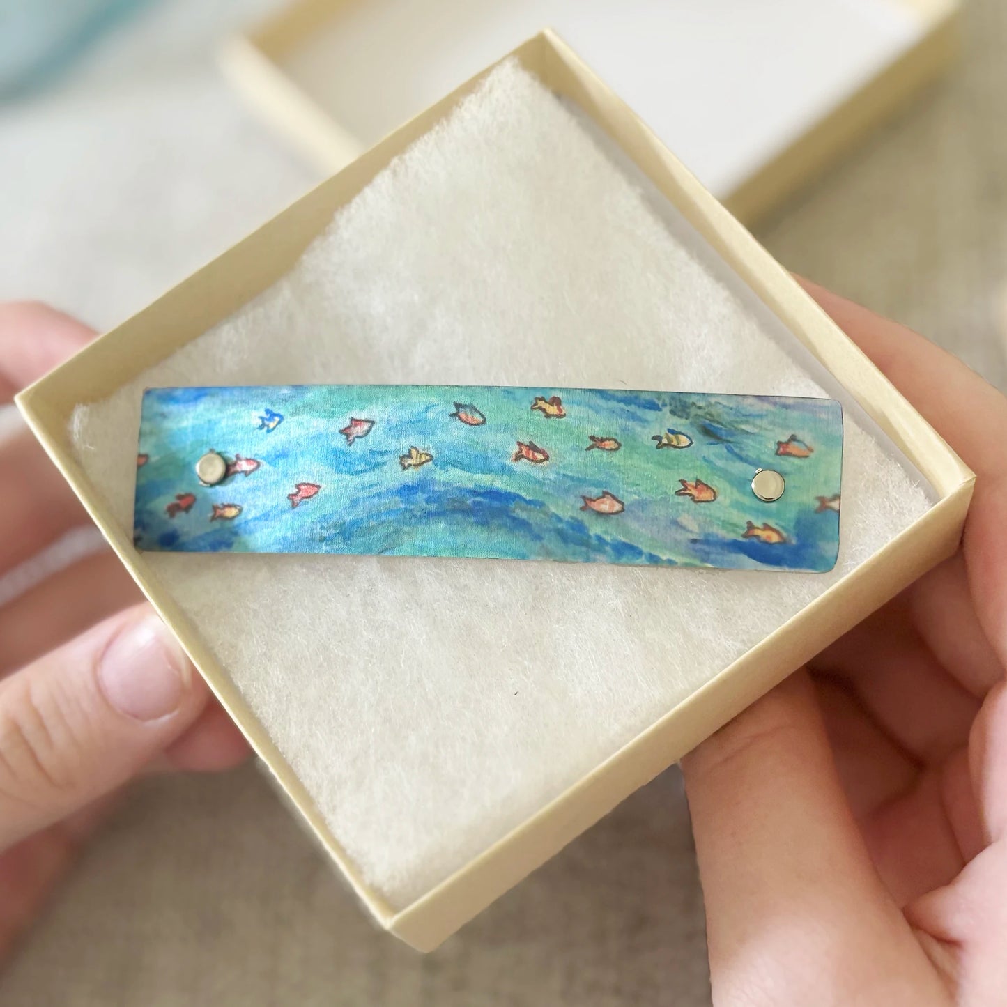 A rectangular barrette with a blue and green swirl wave pattern and small orange fish motifs, displayed inside a cream-colored presentation box held by two hands.