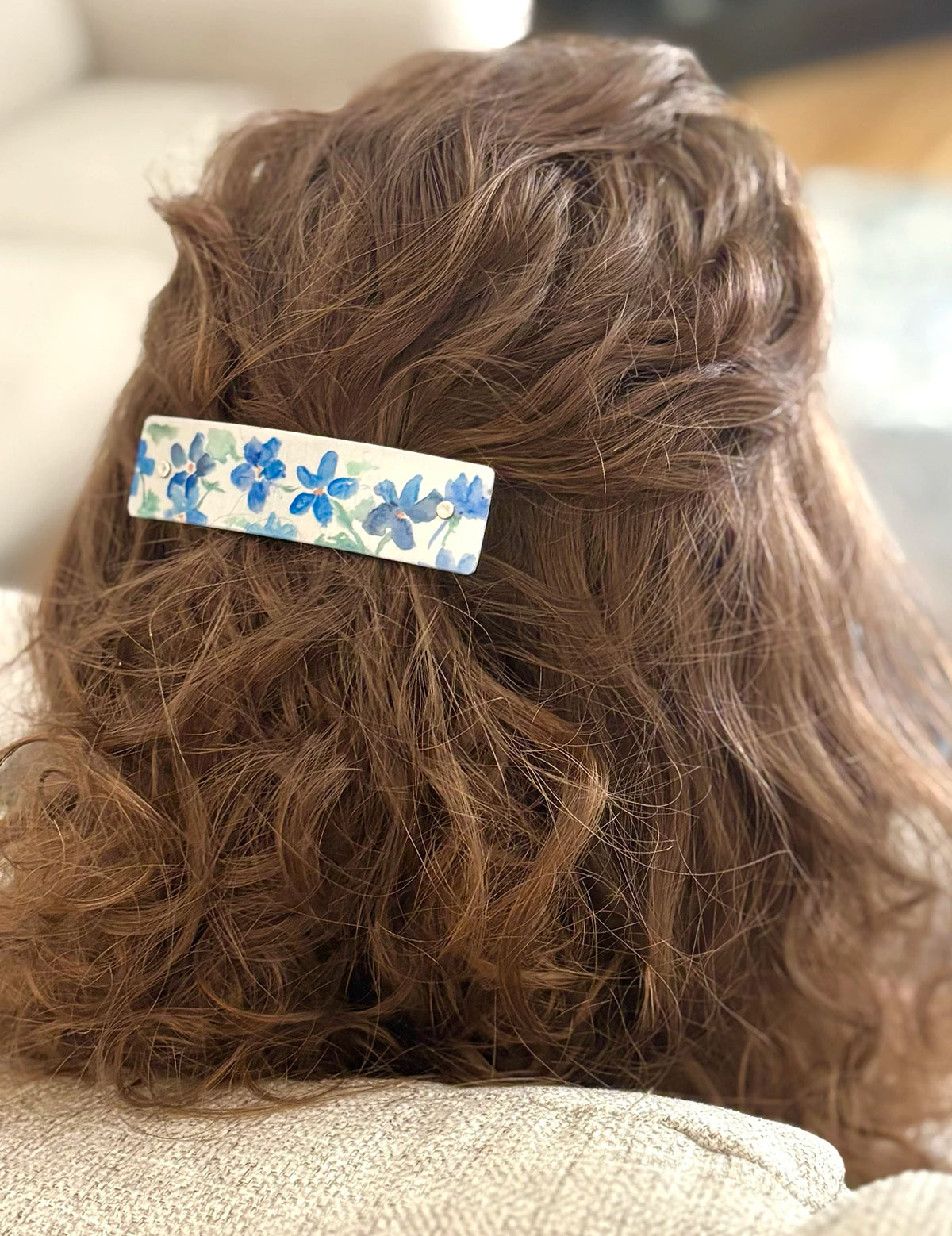 A close-up lifestyle shot of a person's brown hair secured with a rectangular barrette hair clip decorated with a blue floral pattern on a silver background.