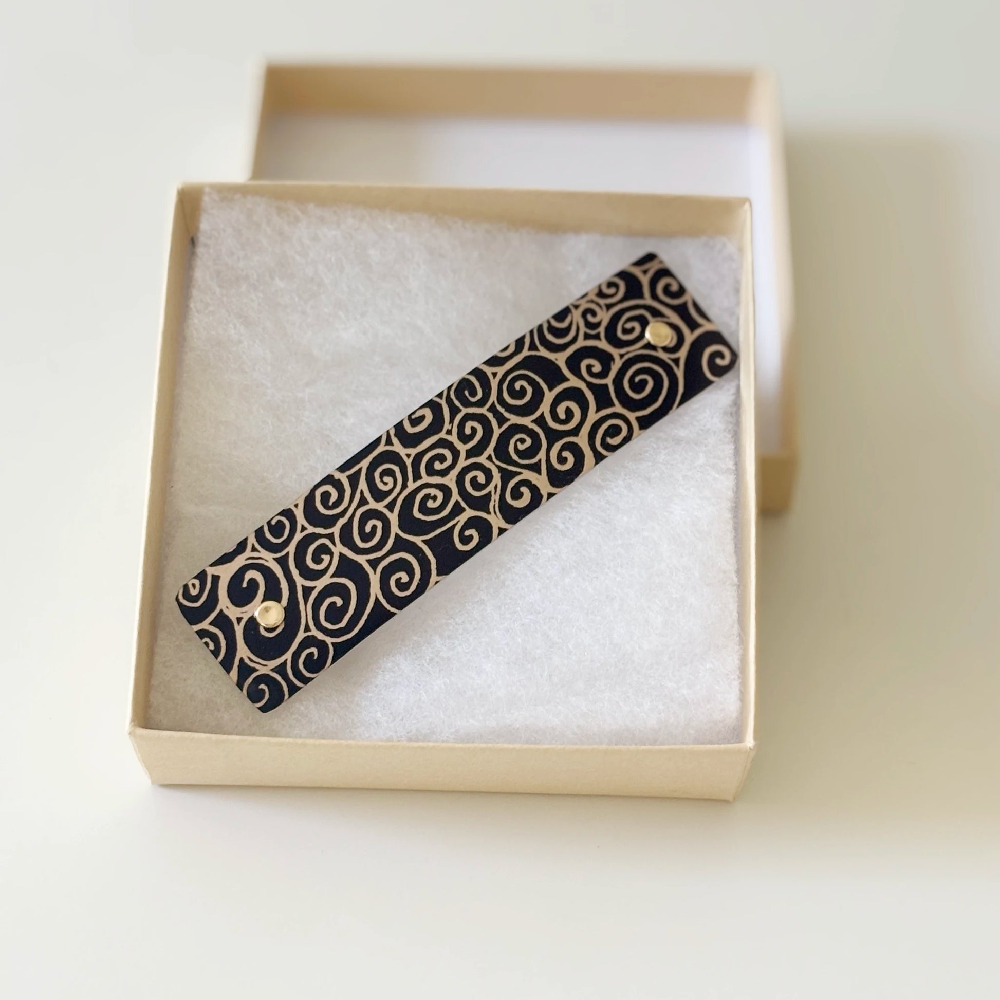 A black metal hair clip with a gold swirl pattern, presented in a cream-colored gift box with white padding, by Robbin V. Davis Studio.