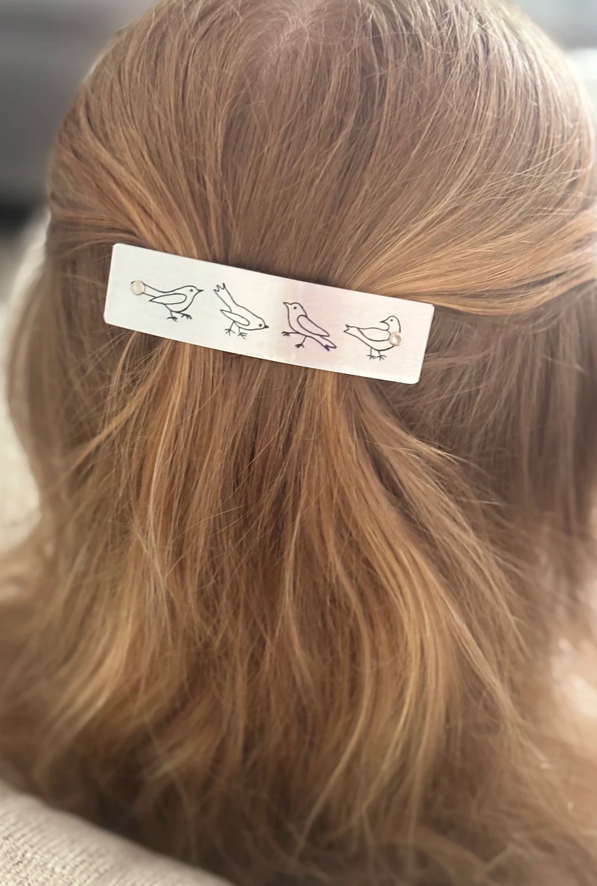 A close-up of a person's light brown hair secured with silver boutique rectangular hair clip decorated with simple black bird illustrations.