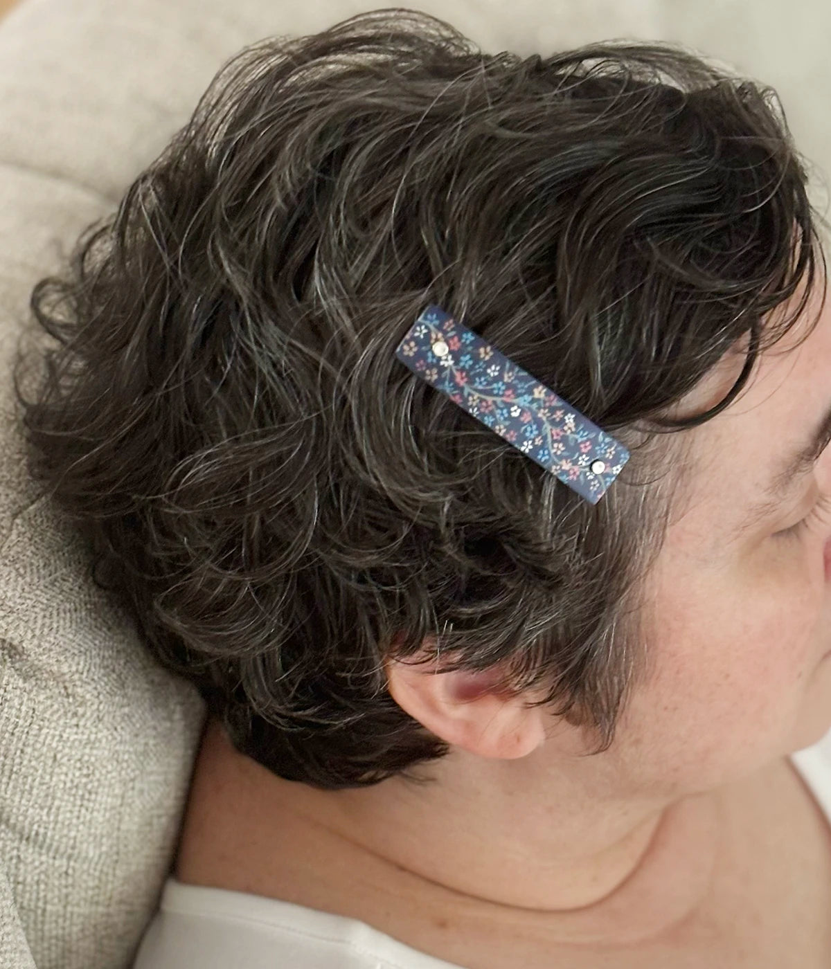 A close-up side view of a person with short curly gray hair wearing a small rectangular hair clip adorned with blue, silver, and red flowers on a navy blue background.