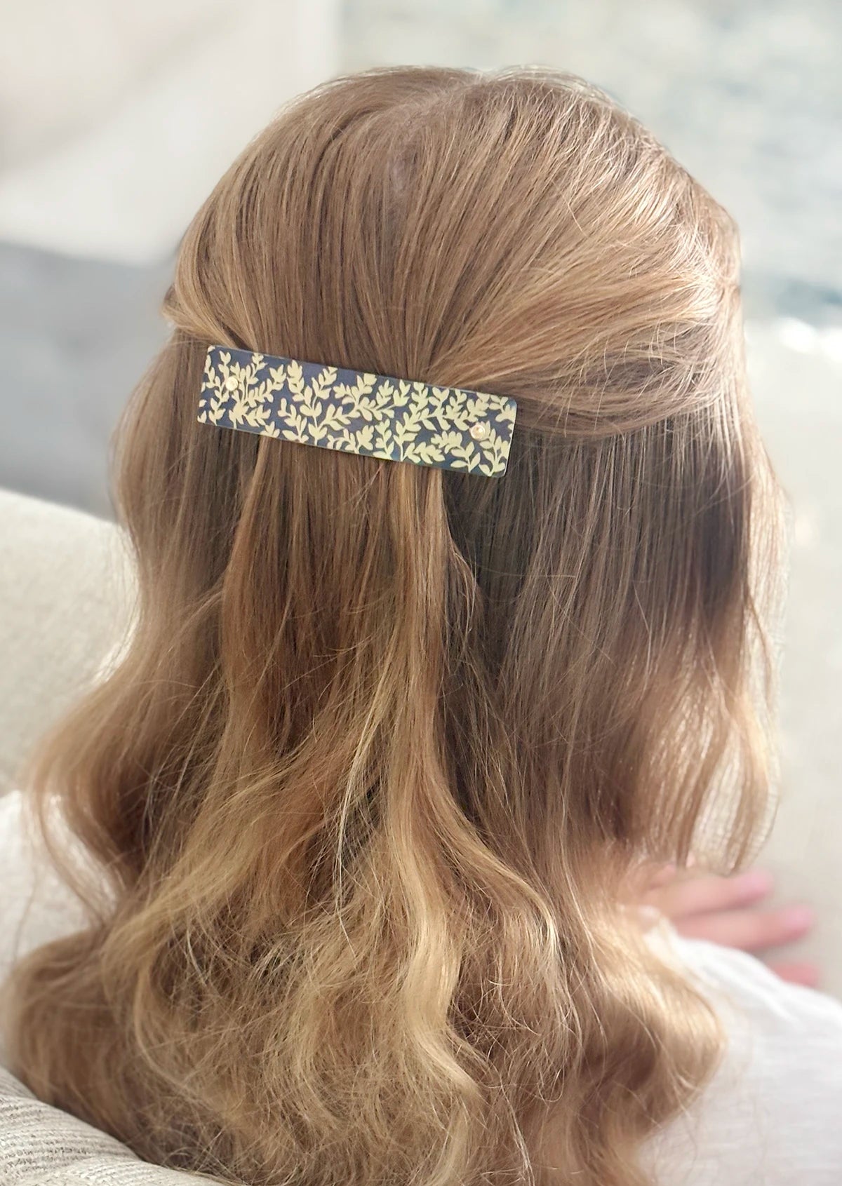 A close-up back view of a person with long, wavy blonde hair wearing a slim rectangular hair clip with a charcoal gray and gold botanical pattern.