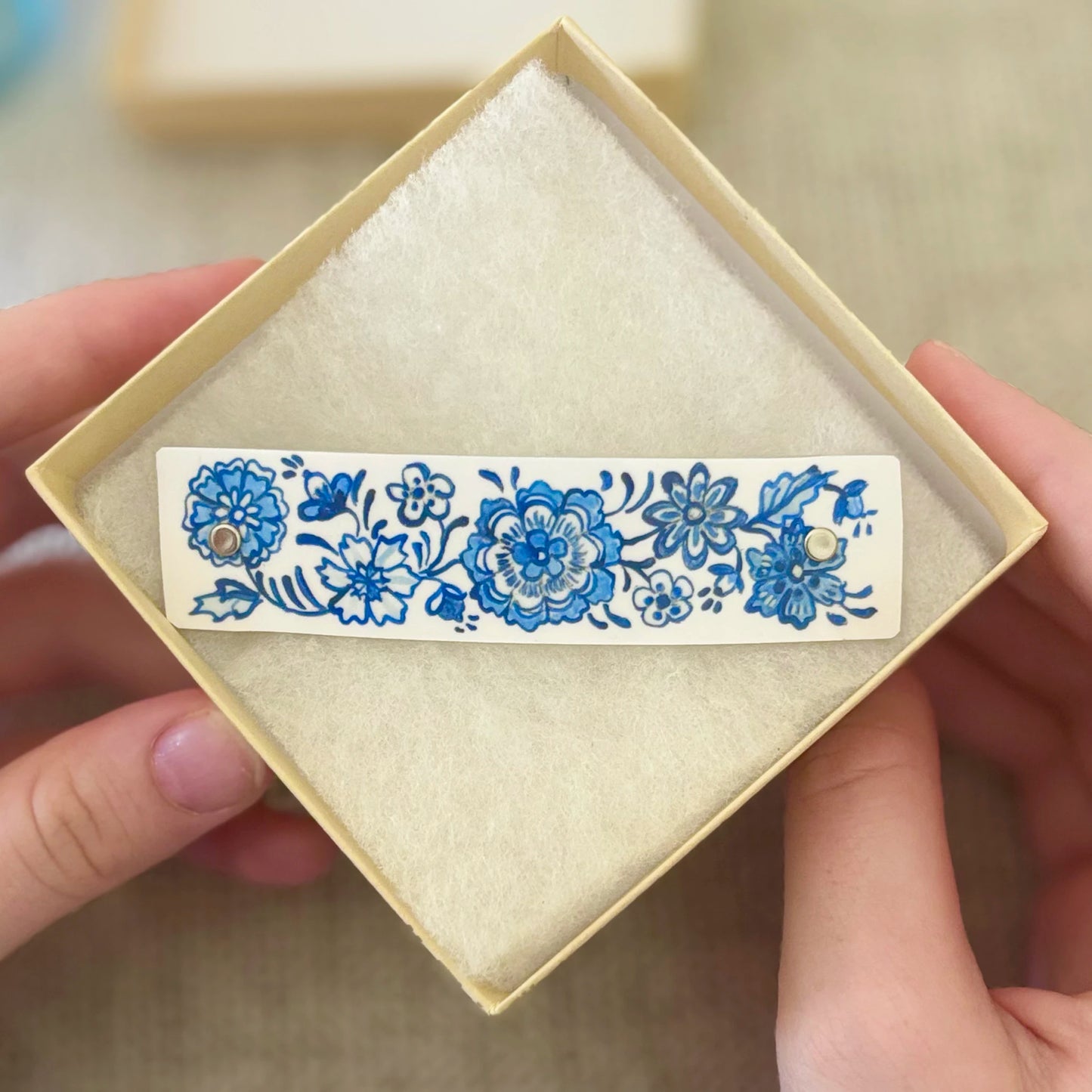 A close-up of a hand holding an open cream-colored jewelry box containing a hair barrette decorated with blue floral patterns, inspired by Delftware.