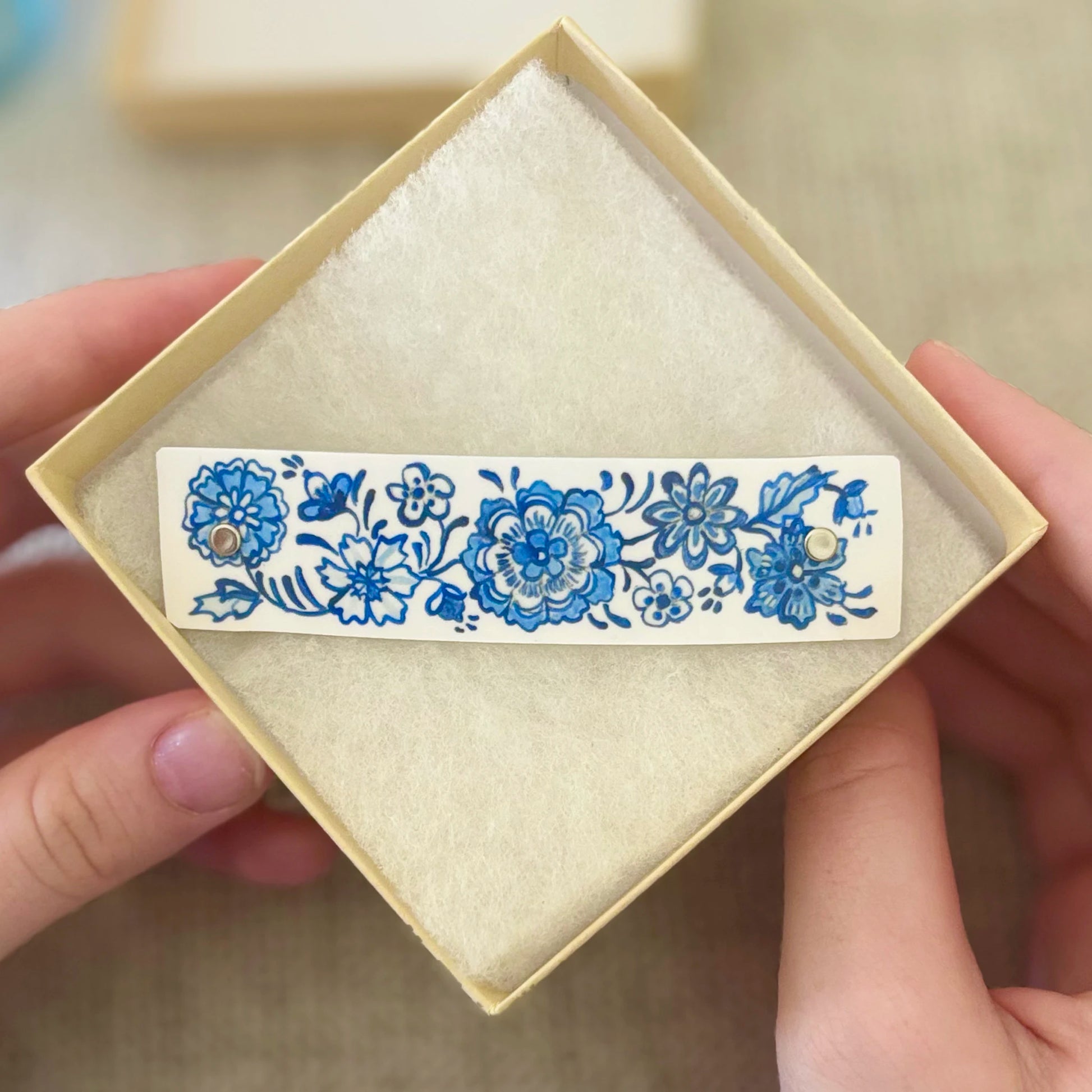 A close-up of a hand holding an open cream-colored jewelry box containing a hair barrette decorated with blue floral patterns, inspired by Delftware.
