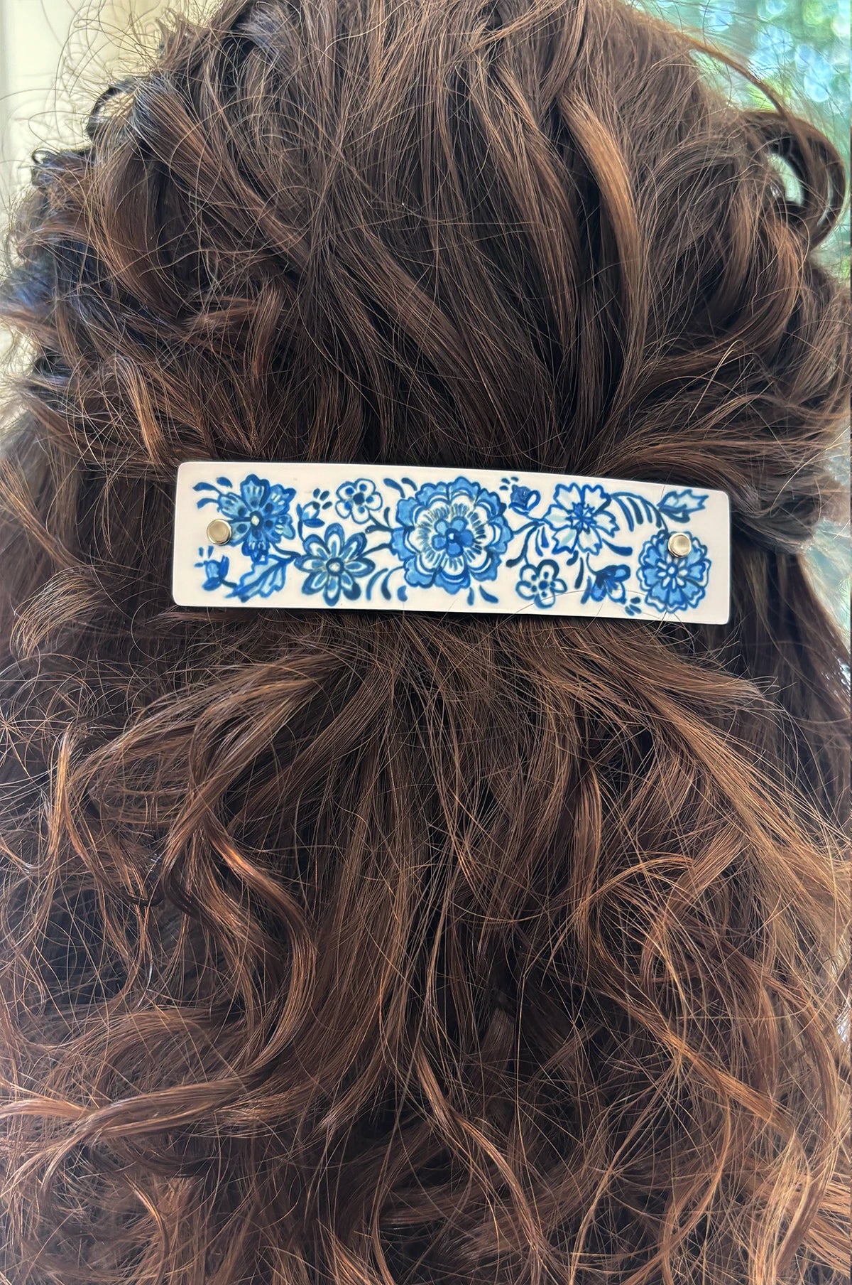 A close-up of a brown wavy hair accessory clip held in place, showing a rectangular white hair barrette with a blue floral pattern.