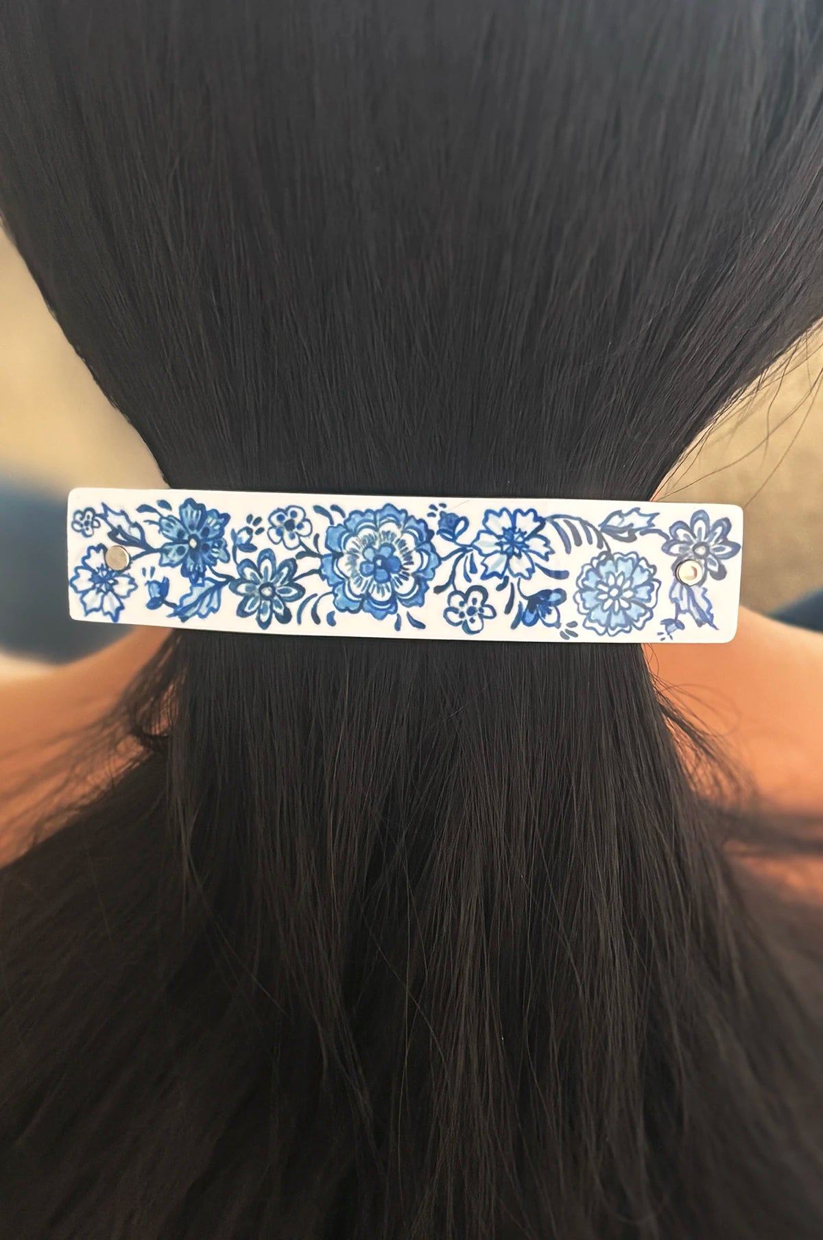 A white boutique hair clip decorated with a blue floral Delftware pattern, shown holding back a section of black hair.