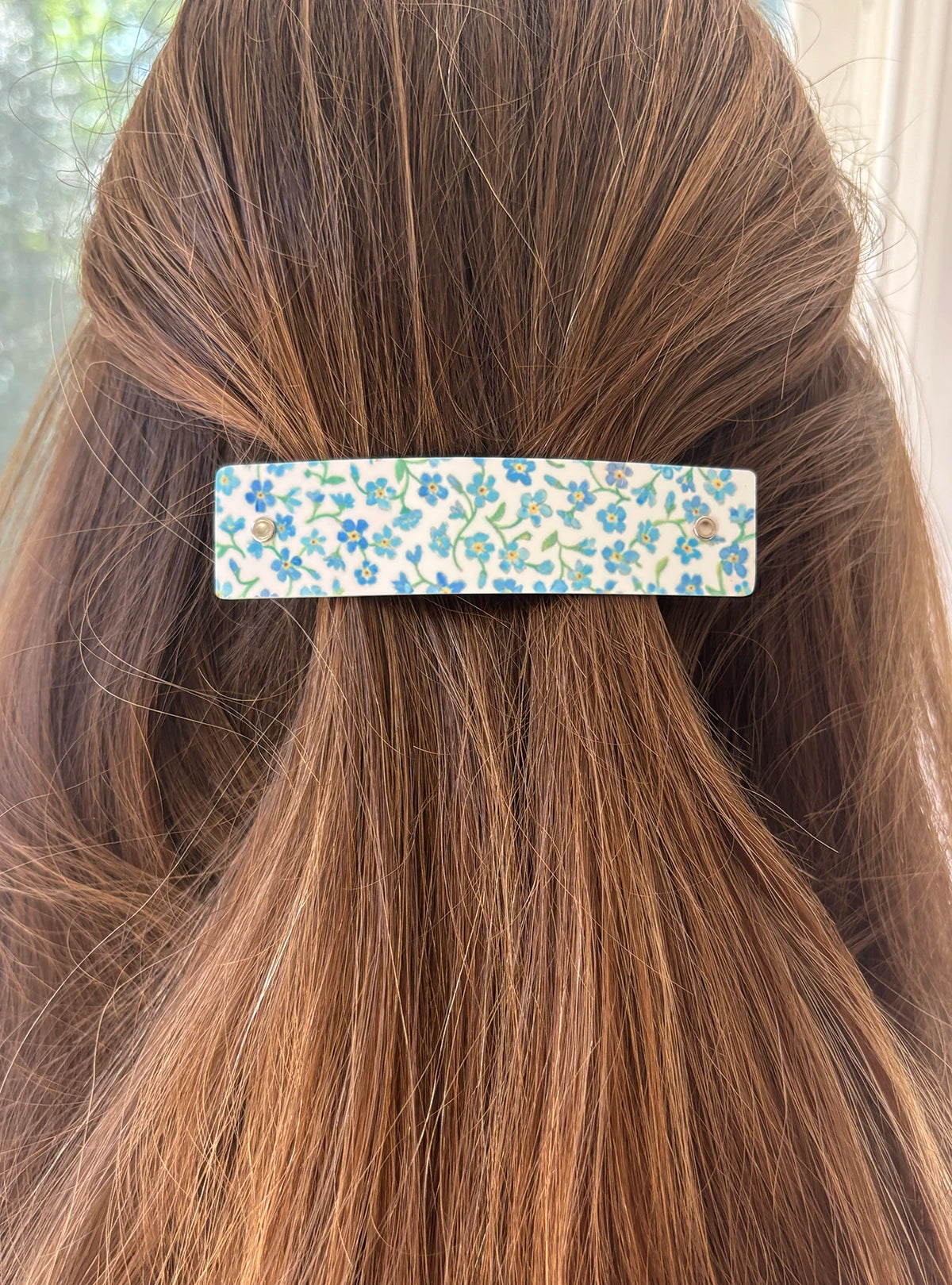 A close-up of a person's brown hair secured with a wide, rectangular hair clip featuring a delicate blue and green floral pattern on a white background.