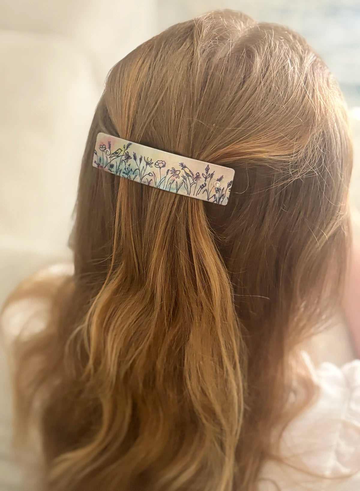 A close-up of a person's wavy blonde hair secured with a slim rectangular hair clip featuring a delicate original floral and bird pattern on a light background.