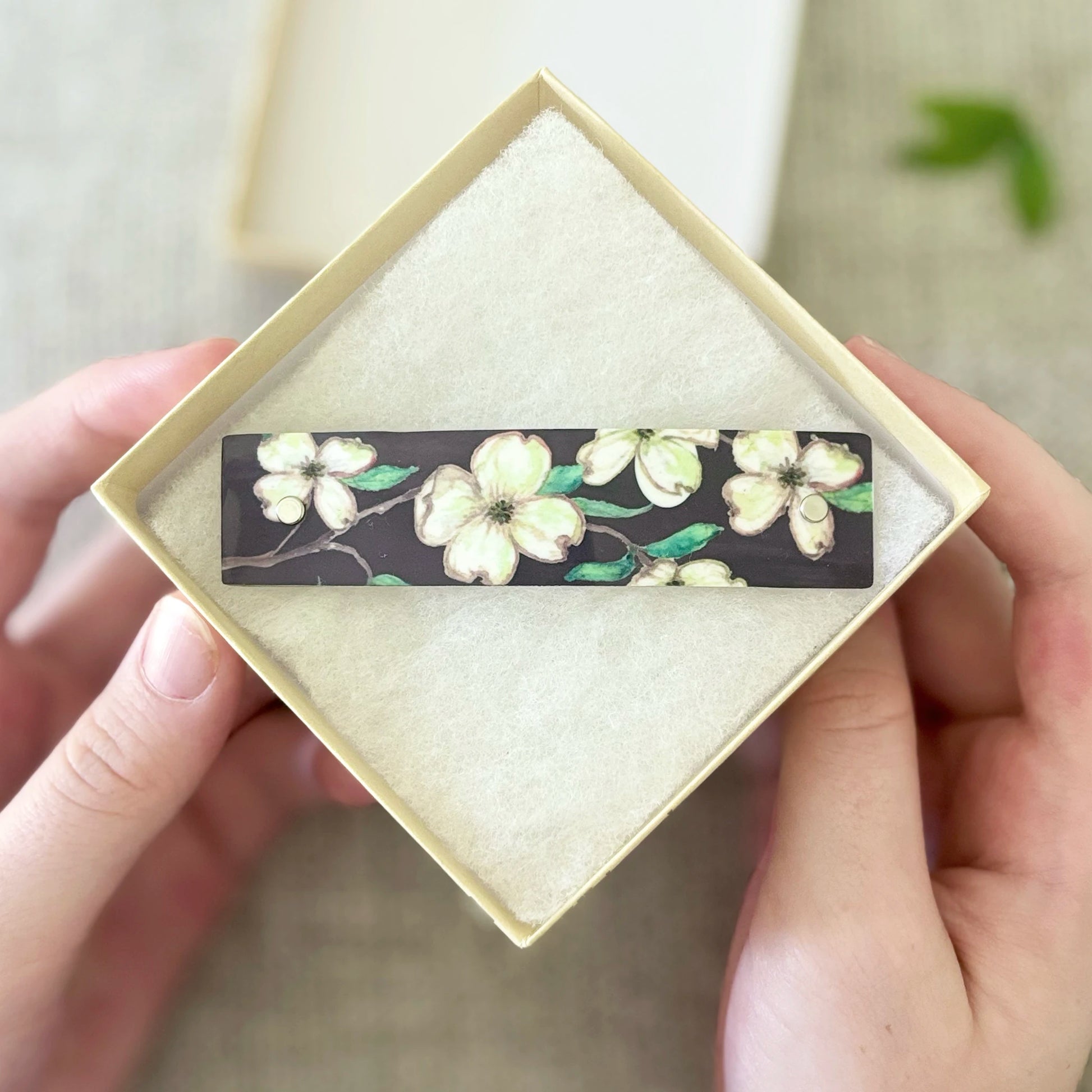 A rectangular barrette with a dark background and white dogwood-style flowers with green leaves, presented in a cream-colored square box held by two hands.