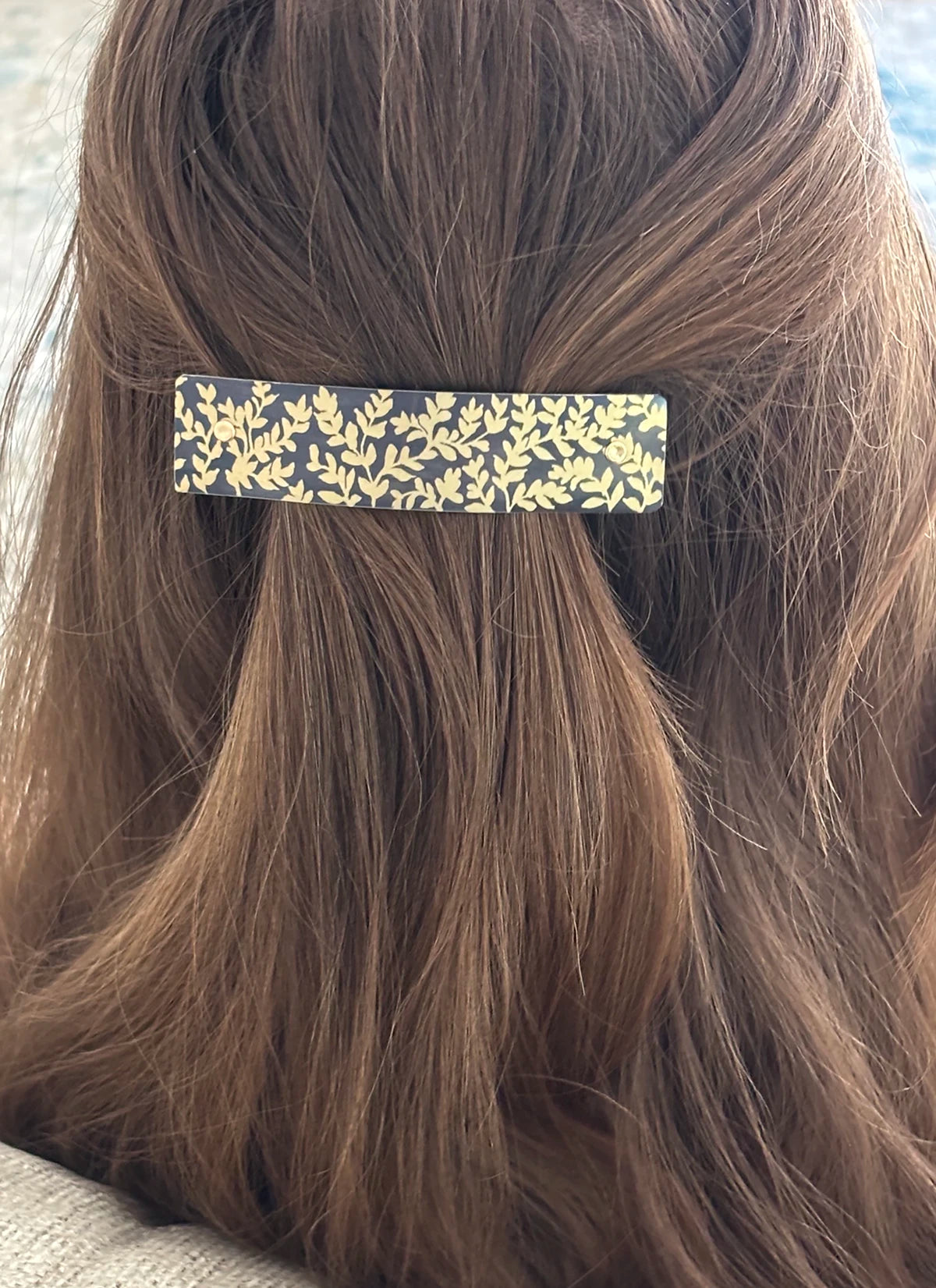 A close-up of a person's brown hair secured with a slim rectangular hair clip featuring a gold leaf pattern on a charcoal gray background.
