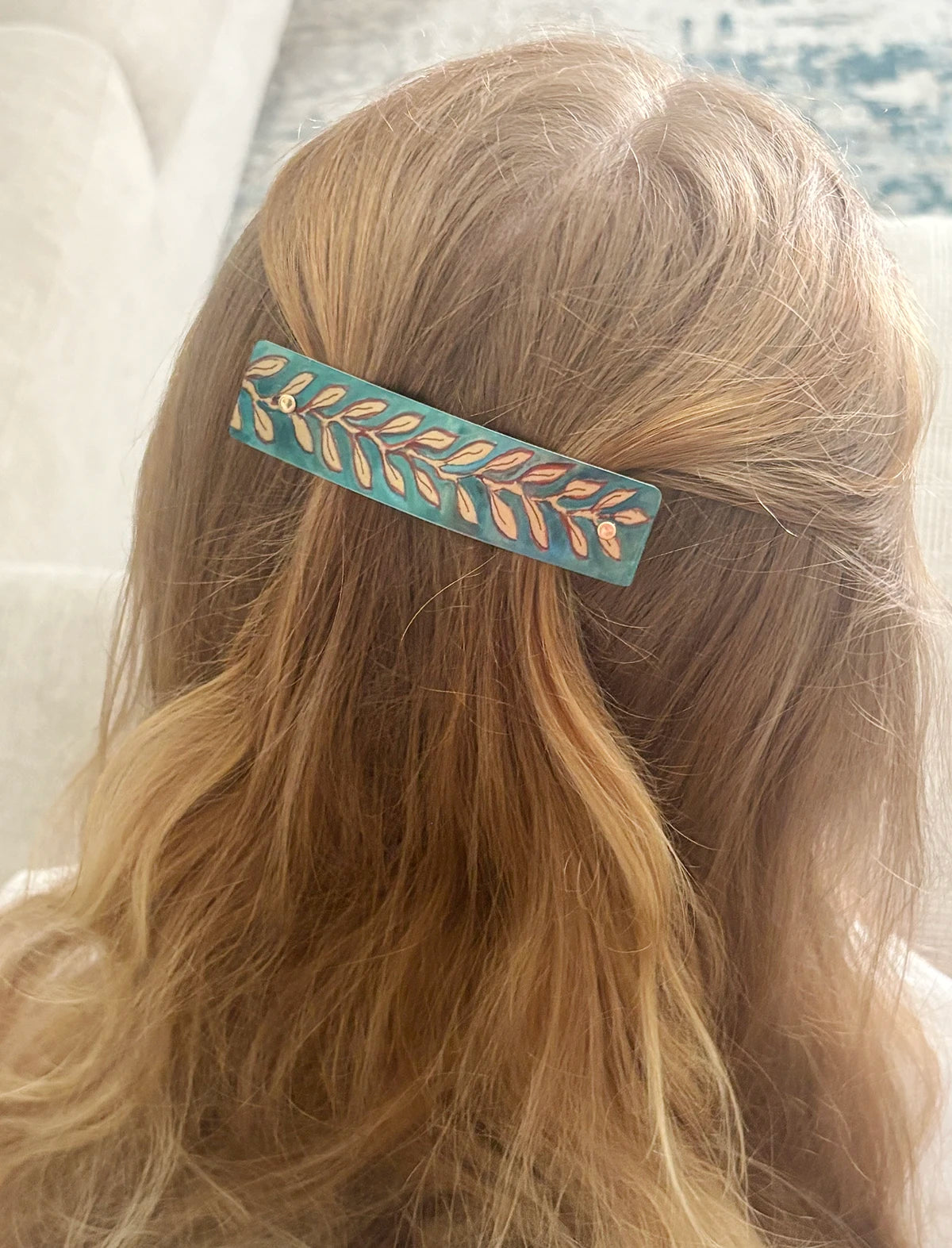 A close-up lifestyle shot of a person's blonde hair secured with a decorative barrette hair clip. The clip has a teal-blue background with a metallic rose-gold leaf pattern.