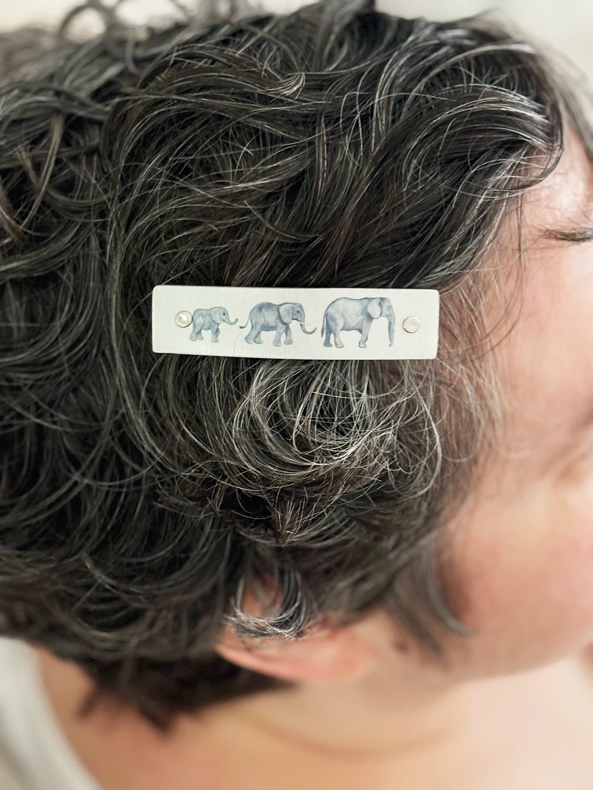 A close-up of a person's dark, curly hair with a silver rectangular hair clip featuring a row of gray elephants.