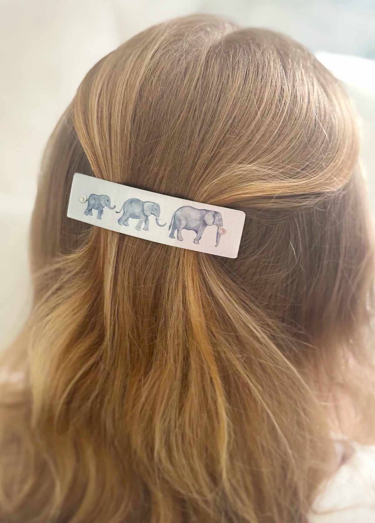 A close-up of a person's blonde hair secured with a silver boutique rectangular hair clip decorated with a watercolor-style illustration of three gray elephants.