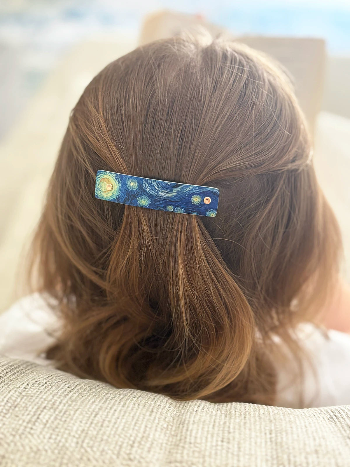 A close-up back view of a person with light brown hair wearing a blue hair clip decorated with a Van Gogh-style starry night pattern.