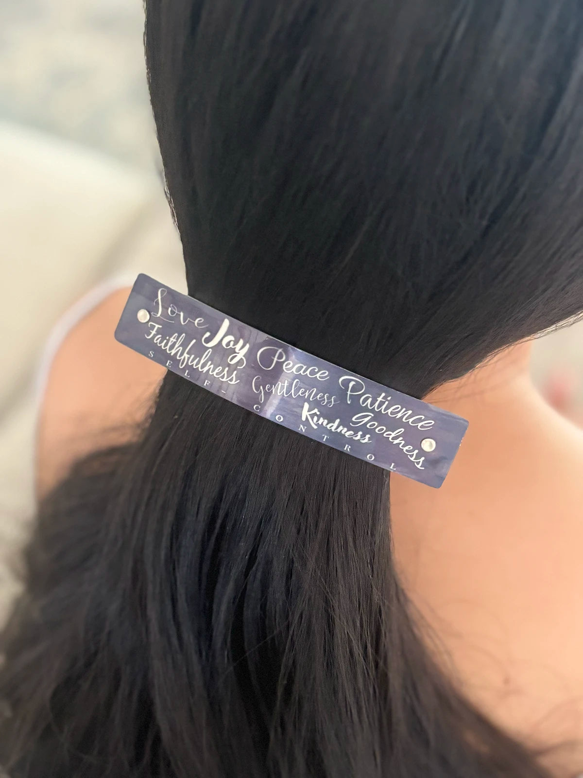 A close-up of a person's dark hair tied back in a low ponytail, featuring a slim, dark gray rectangular hair clip with silver cursive text that reads 'Love Joy Peace Patience Kindness Faithfulness Goodness'.