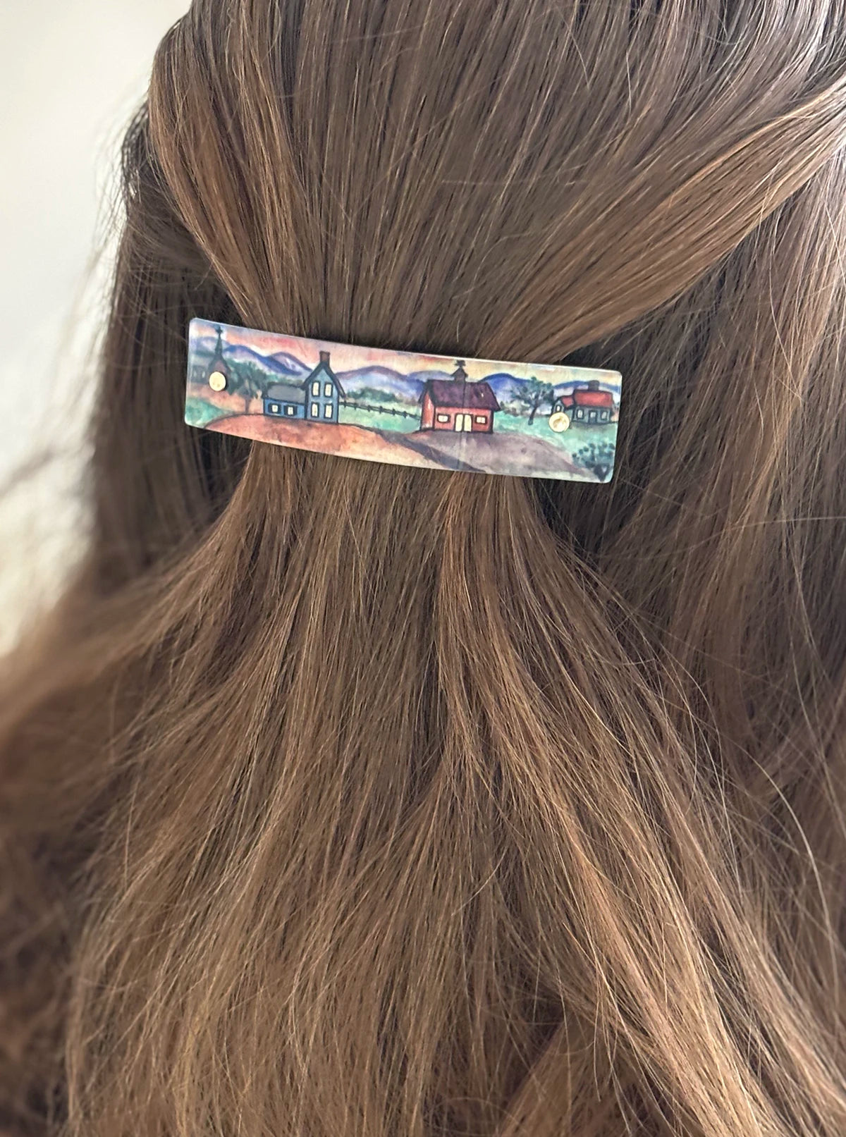 A close-up of a person's brown hair secured with a small rectangular barrette featuring a colorful painted landscape of houses and mountains.