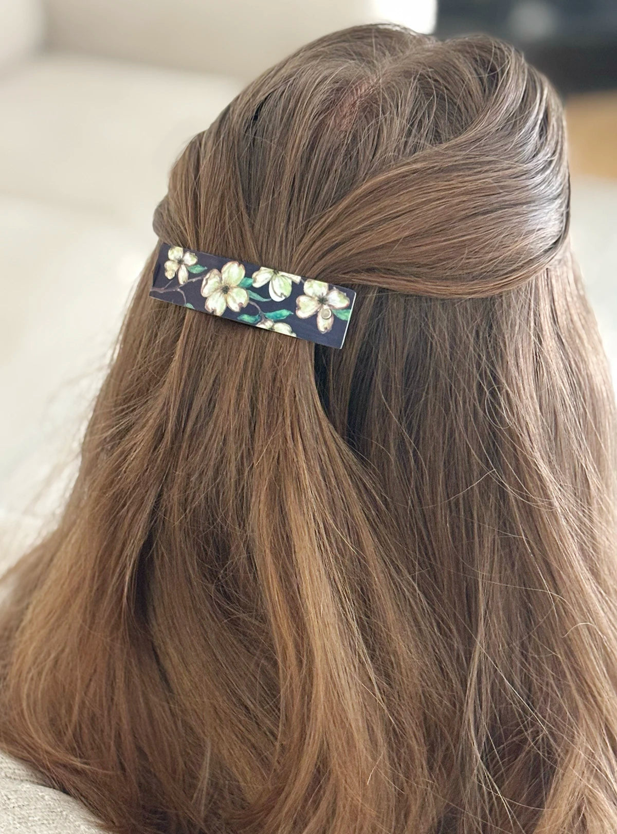 A close-up of a woman's styled brown hair secured with a slim rectangular hair clip featuring a white floral botanical pattern on a dark background.