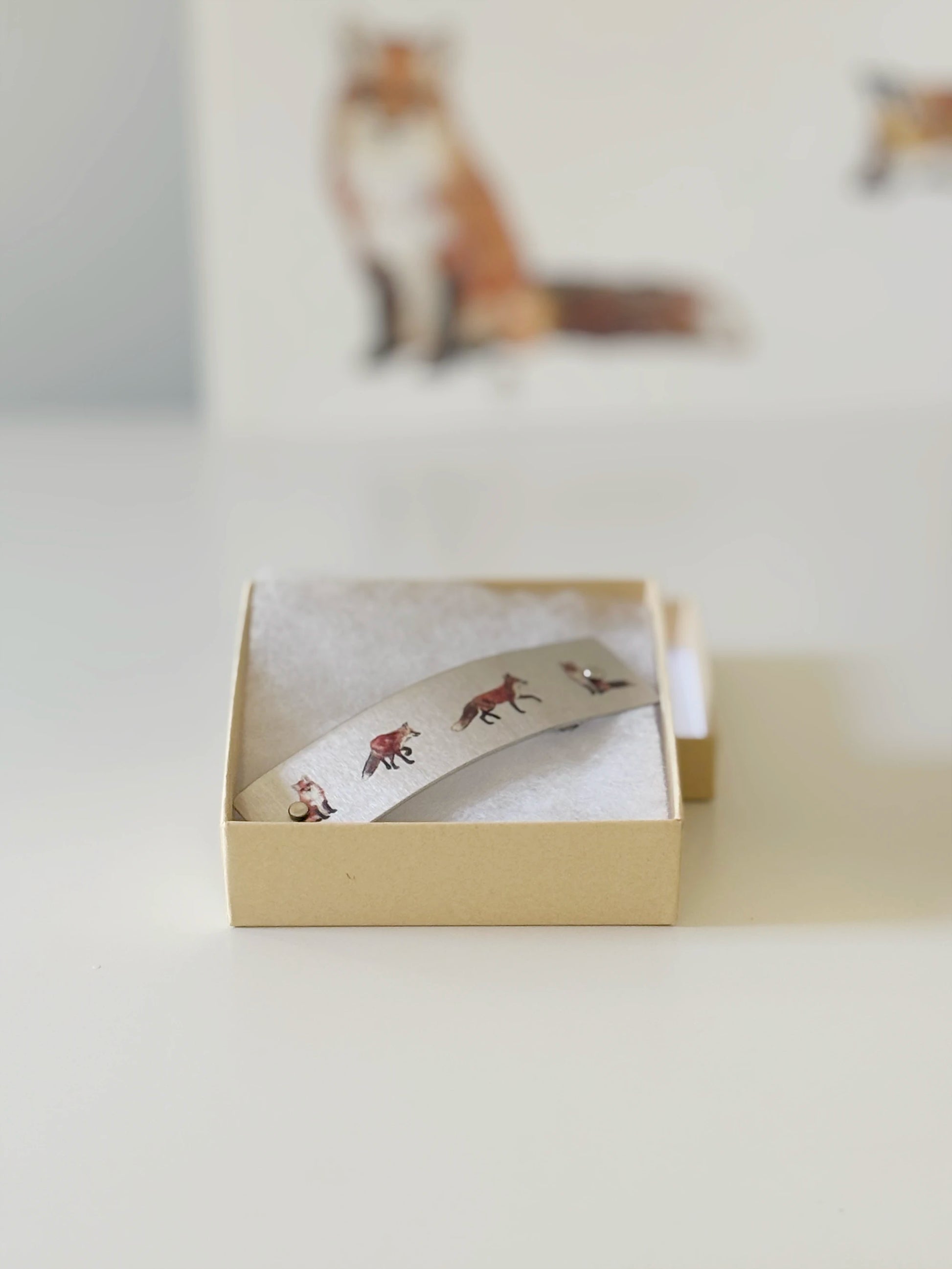 A silver-toned metal hair clip with small watercolor fox motifs, presented in an open beige gift box on a light surface.