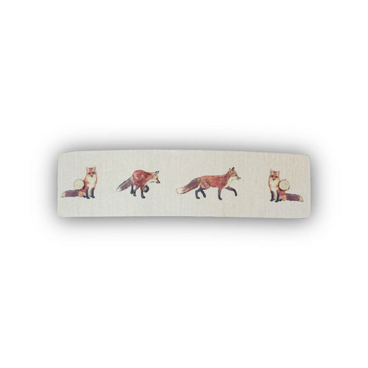 A rectangular hair barrette with rounded corners, featuring a silver metallic background printed with a row of watercolor-style foxes in shades of orange, brown, and cream.