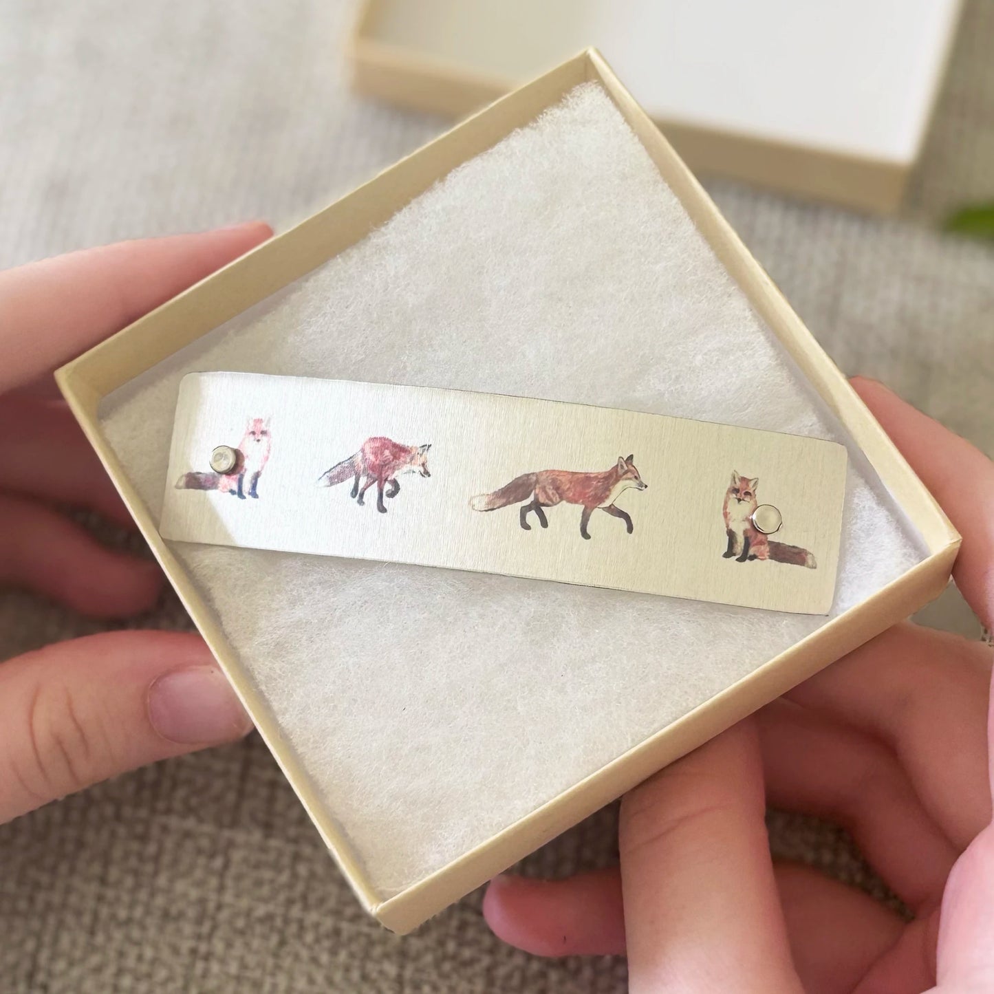 A close-up of hands holding an open beige gift box with a boutique hair barrette featuring illustrated foxes, set against a soft neutral fabric background.