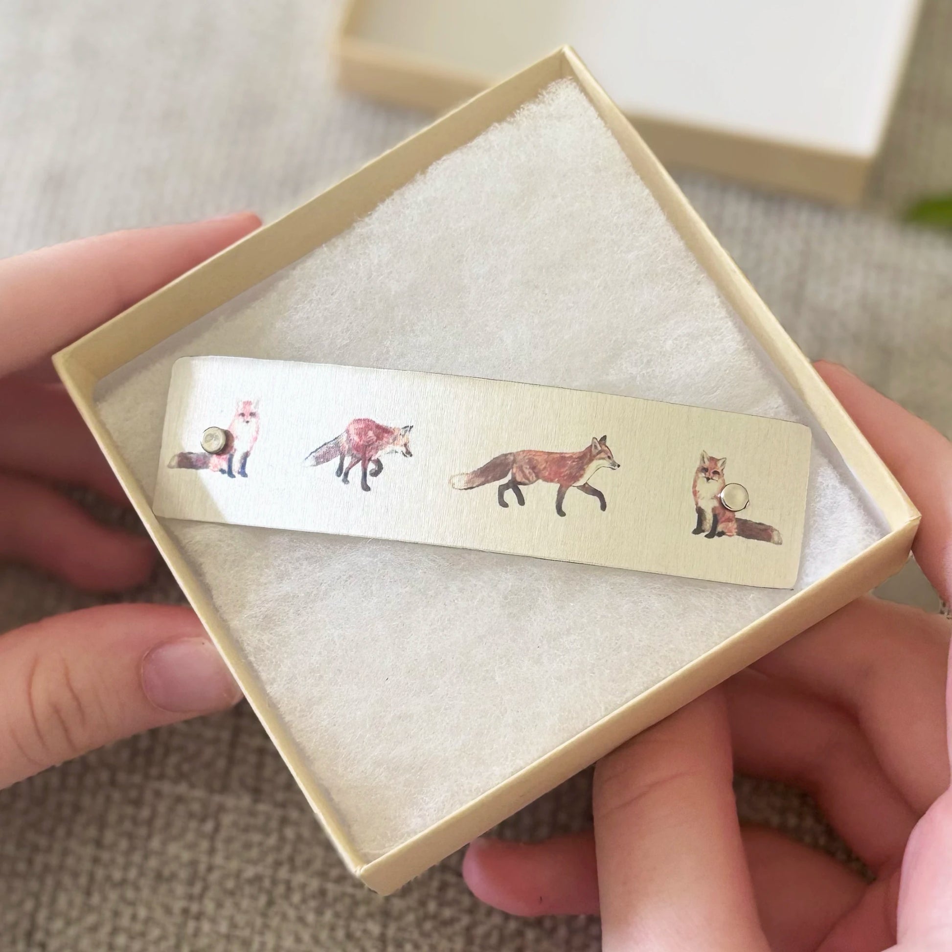 A close-up of hands holding an open beige gift box with a boutique hair barrette featuring illustrated foxes, set against a soft neutral fabric background.