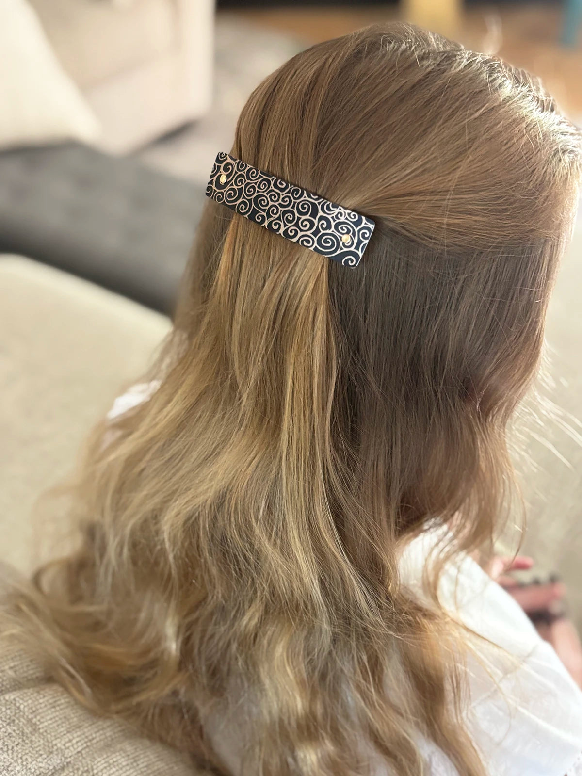 A close-up back view of a person with long blonde hair wearing a decorative black hair clip with gold scrollwork and  gold rivets.