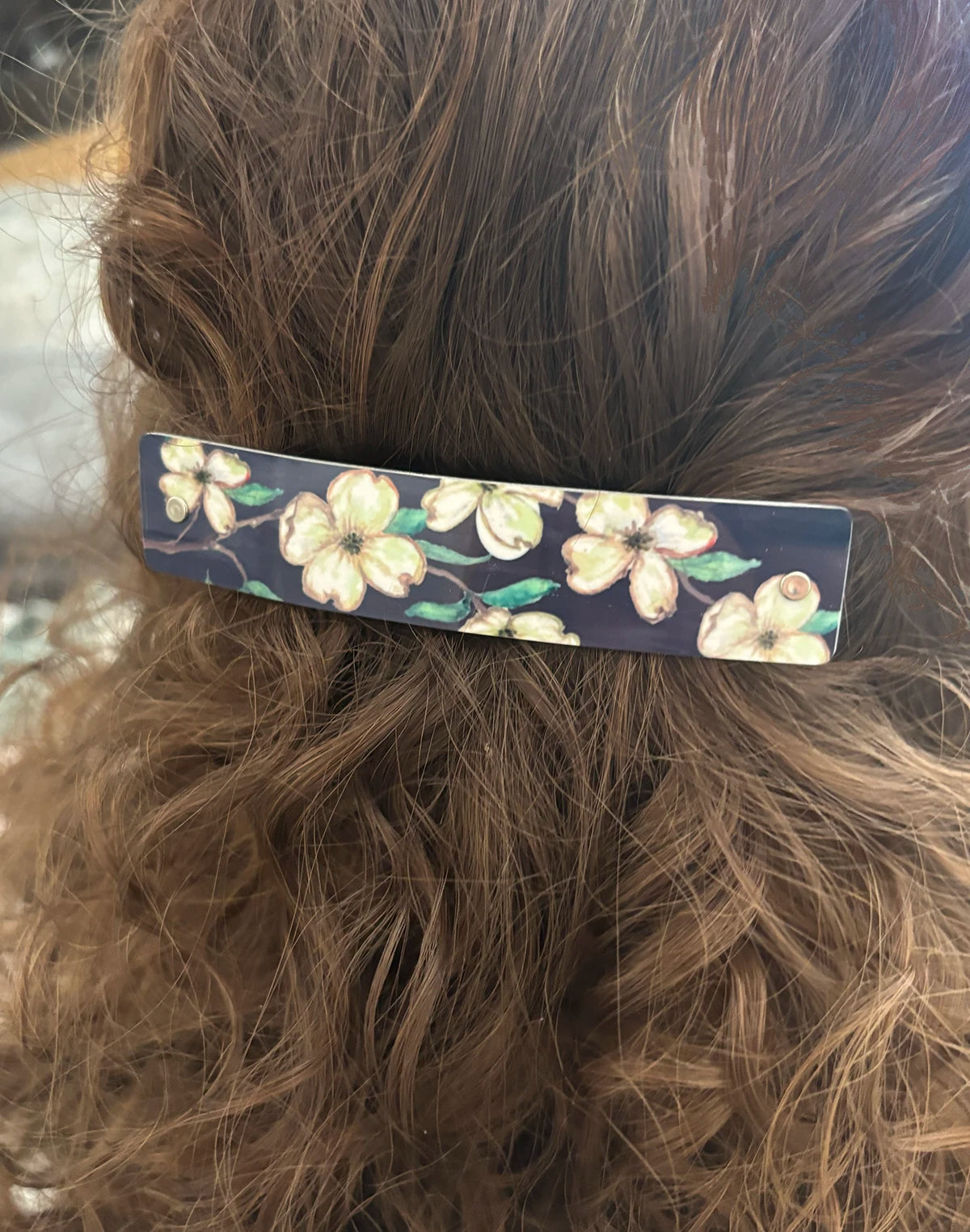 A close-up of brown hair secured with a slim rectangular hair clip featuring a floral pattern of cream blossoms and green leaves on a dark background.