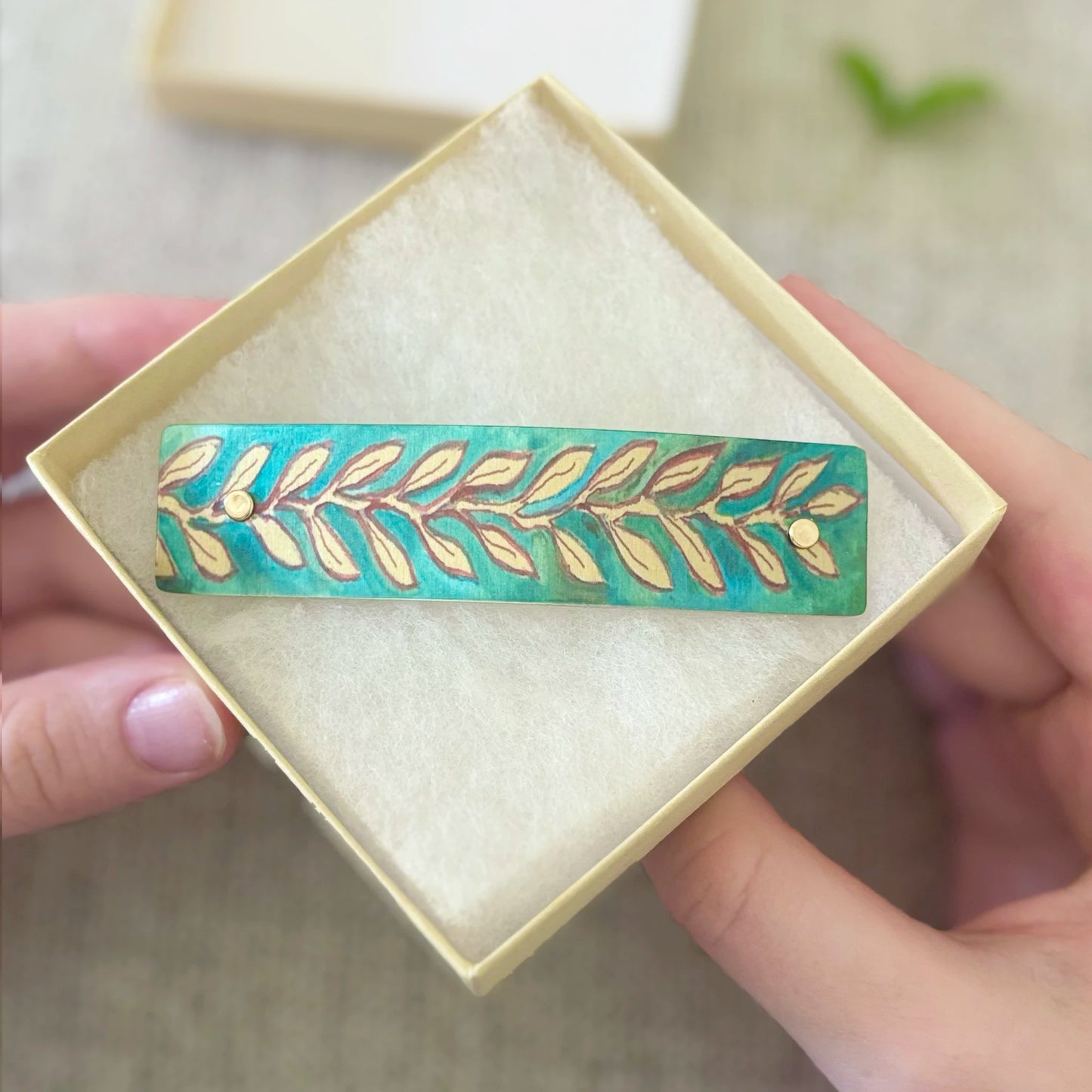 A close-up of a hand holding an open cream-colored jewelry box containing a long barrette with a gold leaf pattern on a turquoise green background.