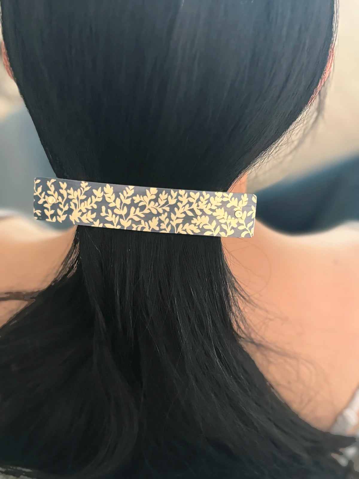 A close-up of a person's black hair secured in a ponytail with a slim rectangular hair clip featuring a gold leaf pattern on a dark background.