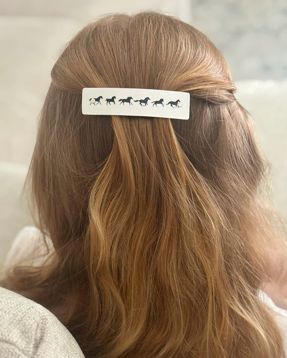 A close-up back view of a person with long strawberry blond hair wearing a silver hair clip decorated with black horse silhouettes.