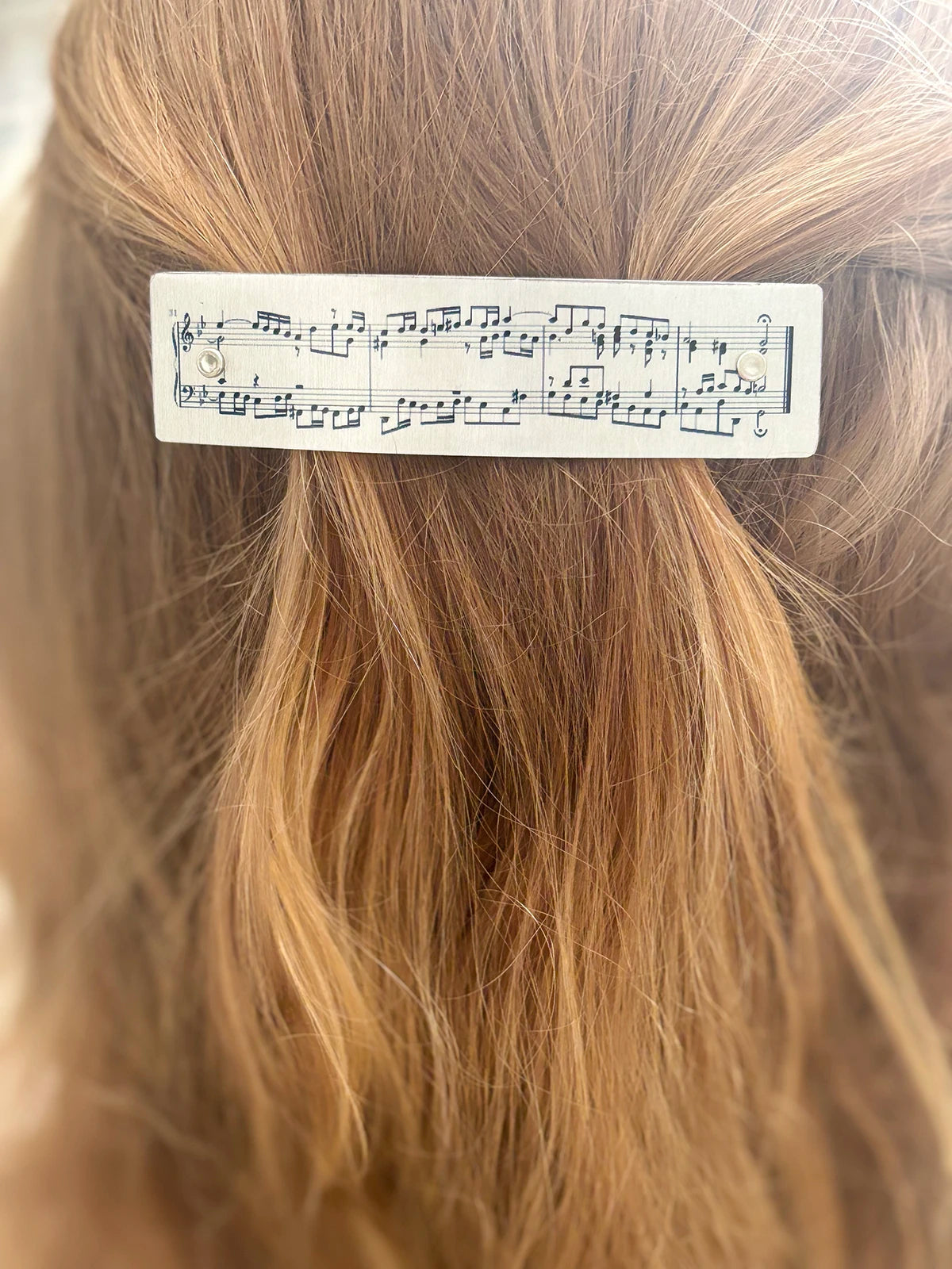 A close-up of strawberry blond hair with a silver handmade rectangular hair clip featuring printed musical notation.