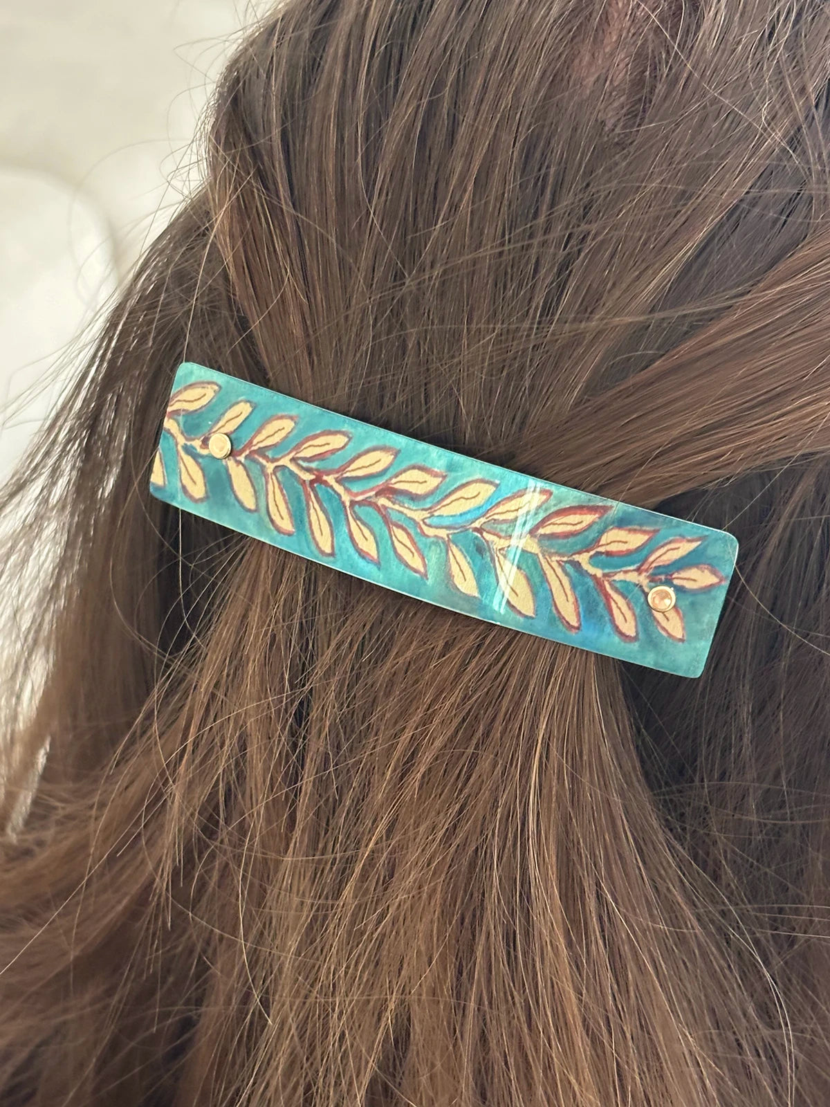 A close-up of a turquoise and teal hair clip with a gold leaf pattern, holding back brown hair.