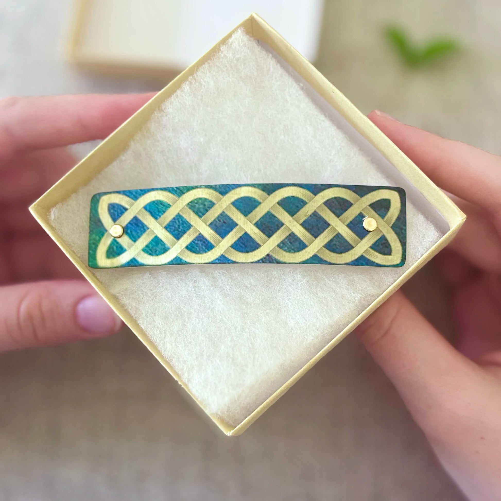 A close-up of a hand holding an open cream-colored jewelry box containing a rectangular barrette with a gold Celtic knot pattern over a textured teal-green background.