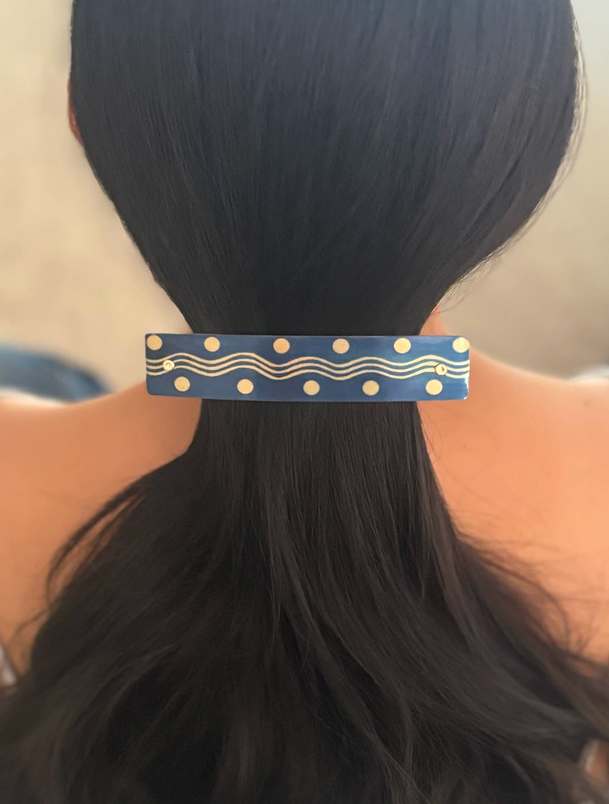 A close-up back view of a person with long black hair secured by a navy blue hair clip decorated with gold polka dots and wavy lines, set against a soft beige background.