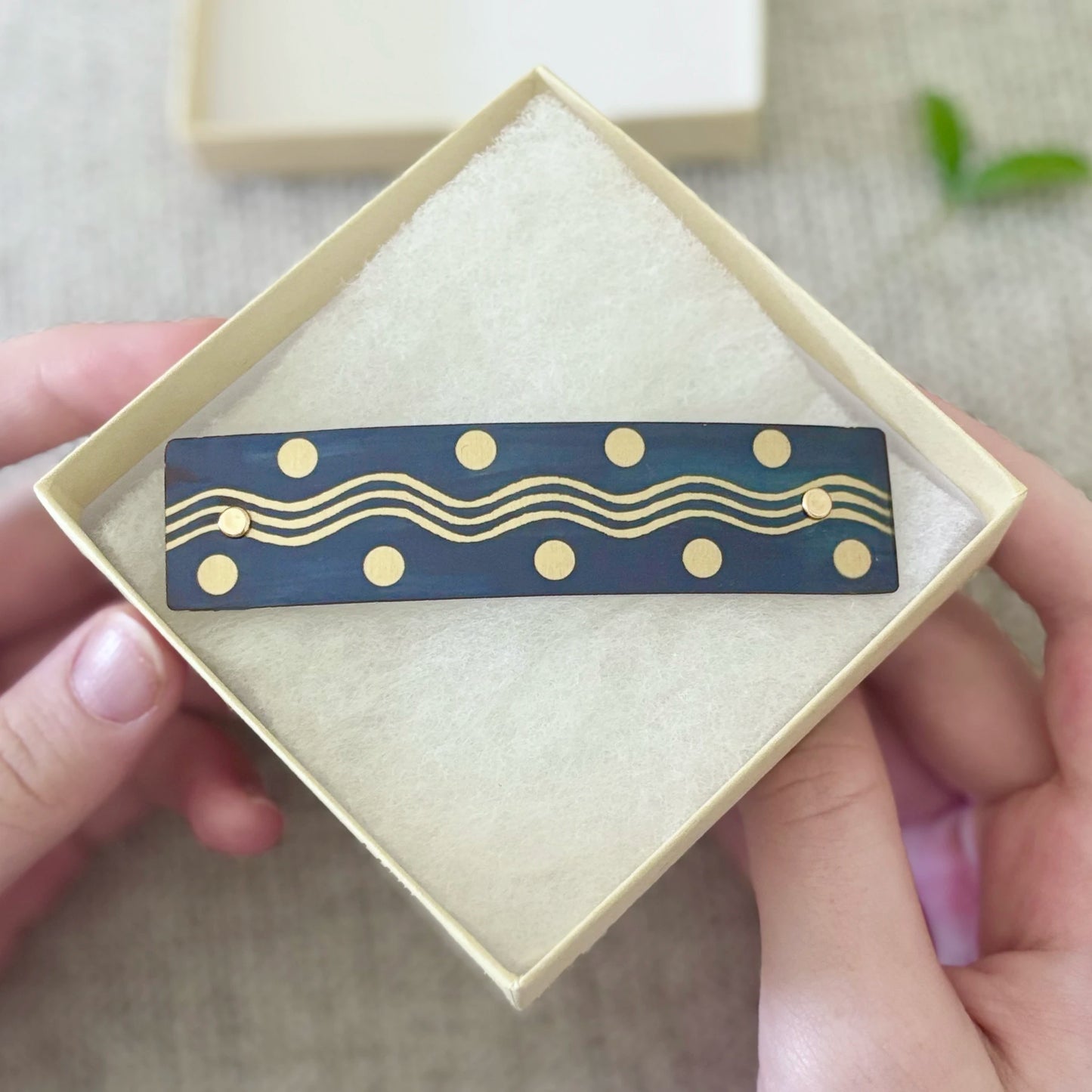 A rectangular navy blue hair barrette with gold wavy lines and dots, presented in an open cream-colored gift box held by hands.