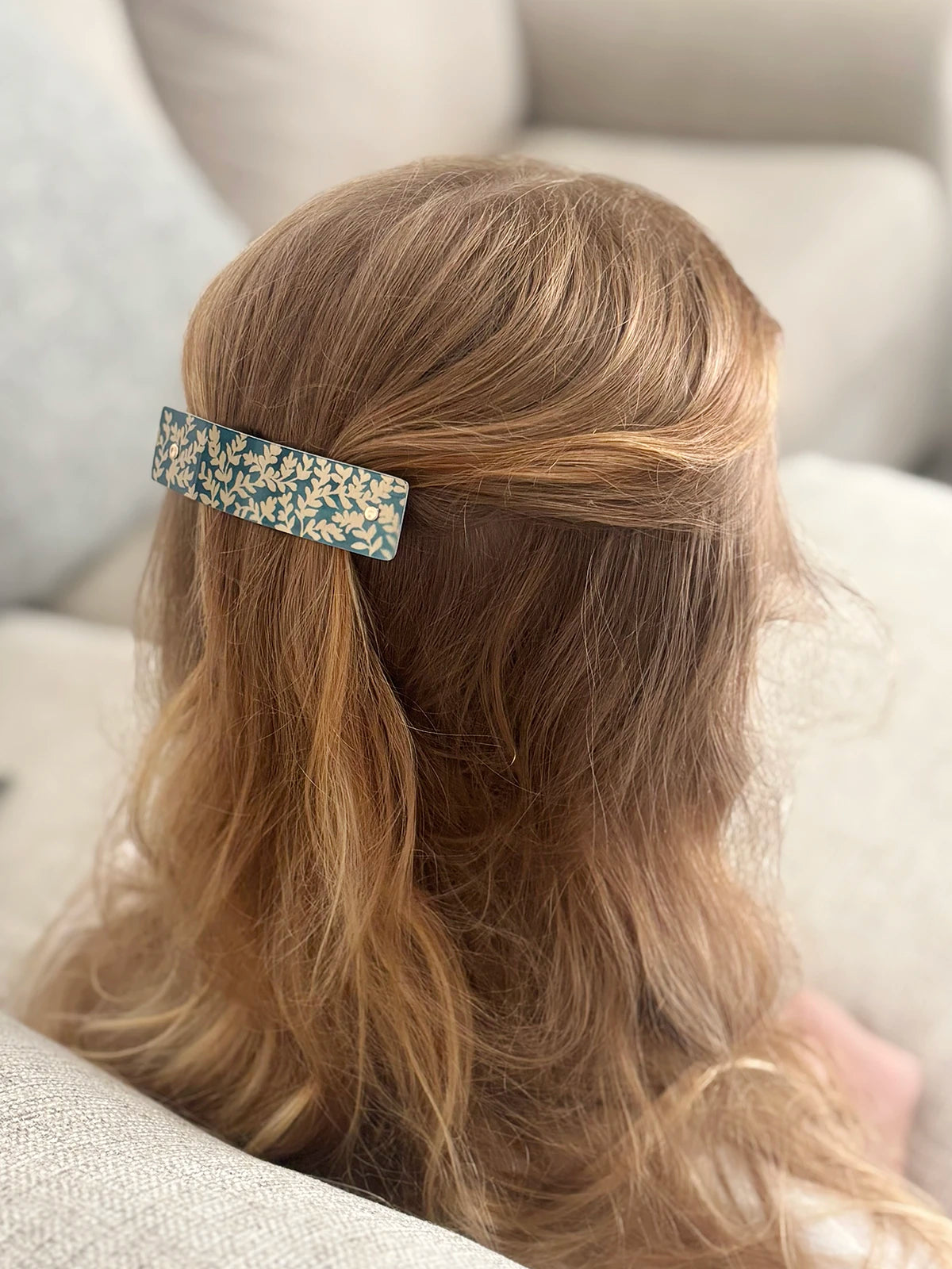 A close-up lifestyle shot of a person with long, wavy strawberry blond hair wearing a rectangular hair clip. The clip features an artistic green and gold botanical pattern.