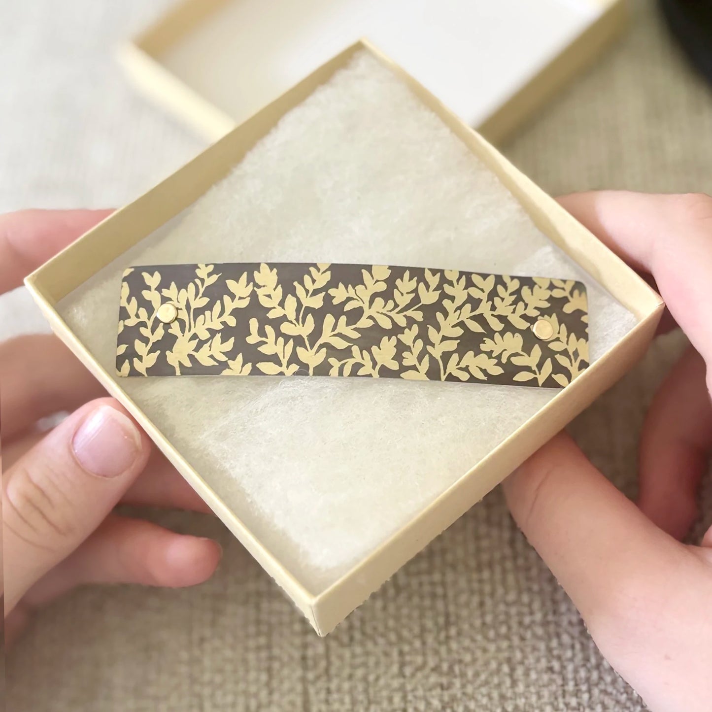 A close-up of a hand holding a square cream-colored gift box containing a slim rectangular hair clip with a repeating gold leaf pattern on a charcoal background.