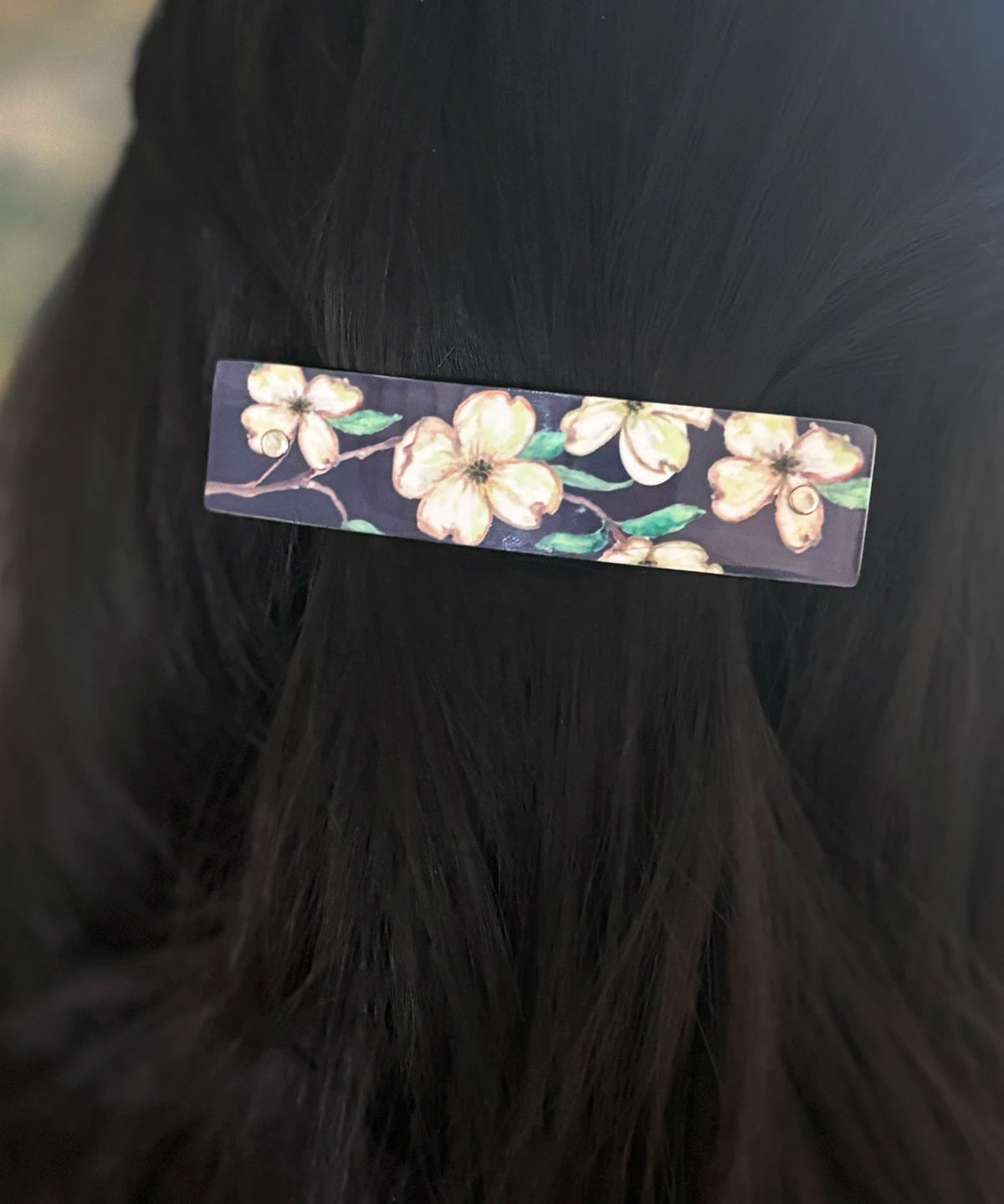 A rectangular hair barrette with a floral print of cream-colored dogwood blossoms and green leaves on a dark background, shown clipped to a section of black hair.