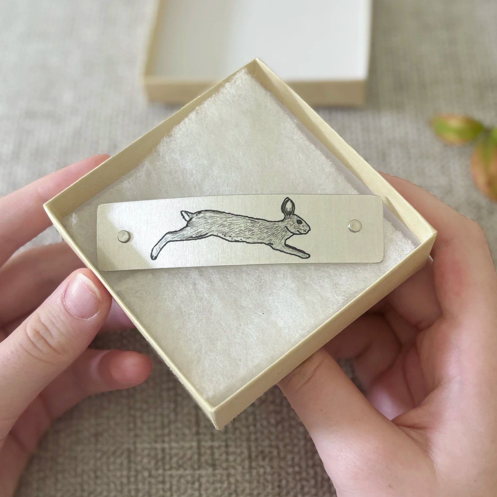 A silver metal barrette adorned with a leaping rabbit, held in a cream-colored square box with a soft cream insert, held by two hands against a neutral textured background.